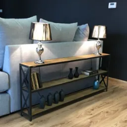 Metalowa konsola z drewnianymi półkami w stylu industrialnym, ustawiona za szarą sofą, z czarnymi wazonami i książkami na półkach, oświetlona dwiema lampkami nocnymi z czarnymi abażurami.
