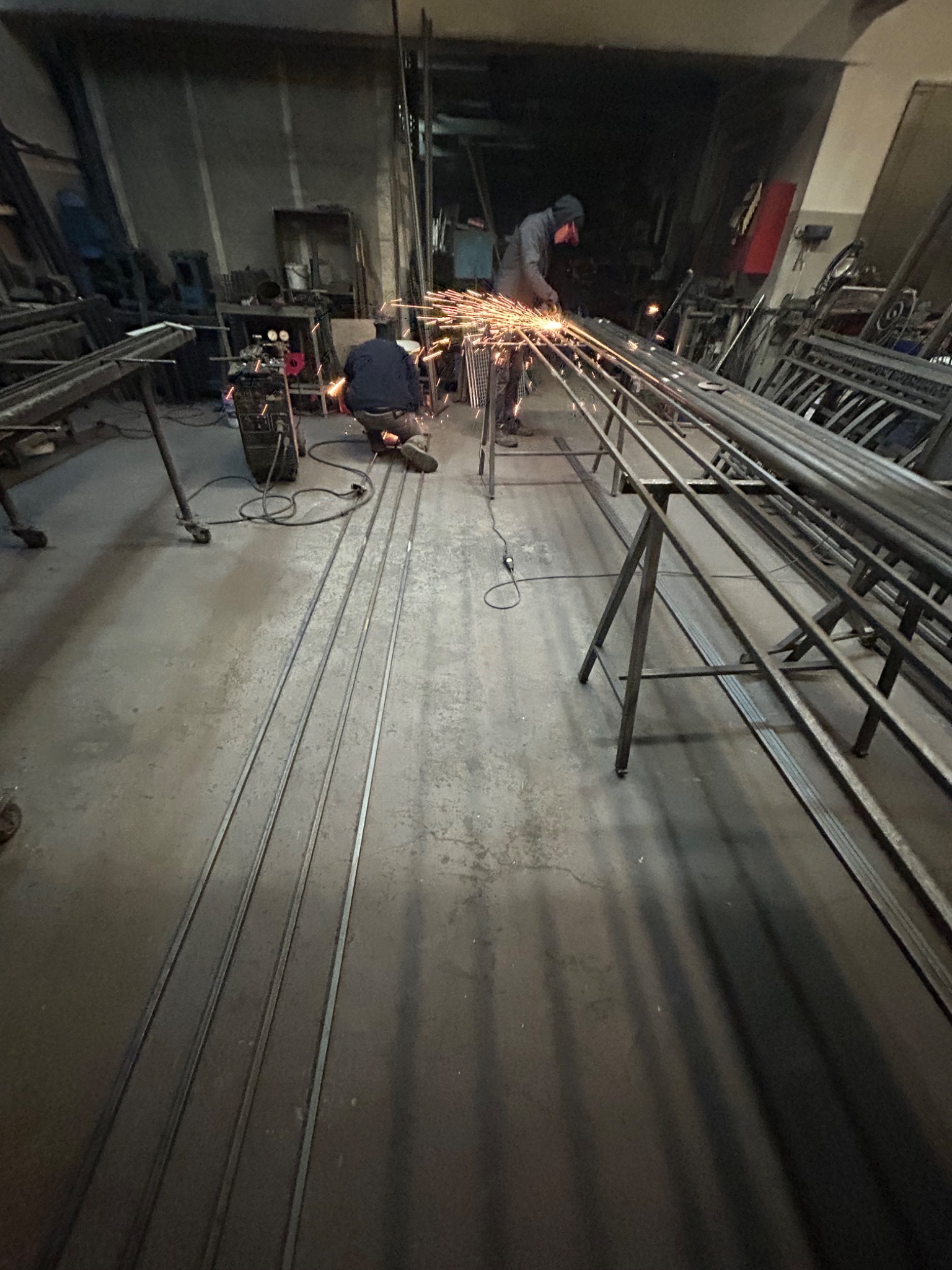 Spawanie konstrukcji metalowej w warsztacie: dwóch spawaczy pracuje nad długimi, metalowymi elementami, iskry latają, widoczny sprzęt spawalniczy i surowe otoczenie.