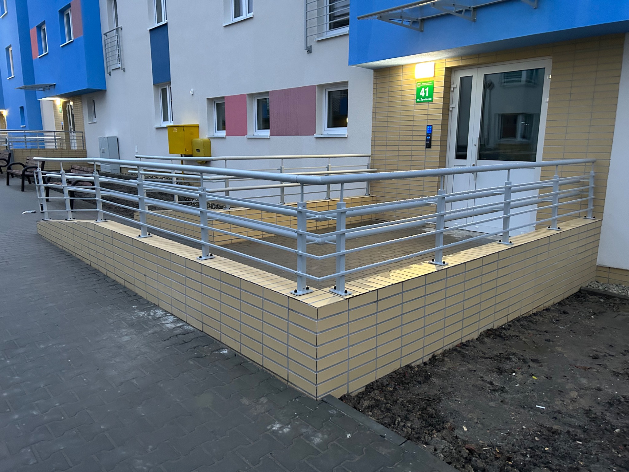 Zewnętrzna balustrada ze stali nierdzewnej, zabezpieczająca podjazd dla osób niepełnosprawnych przy wejściu do budynku mieszkalnego, z numerem 41 nad drzwiami.