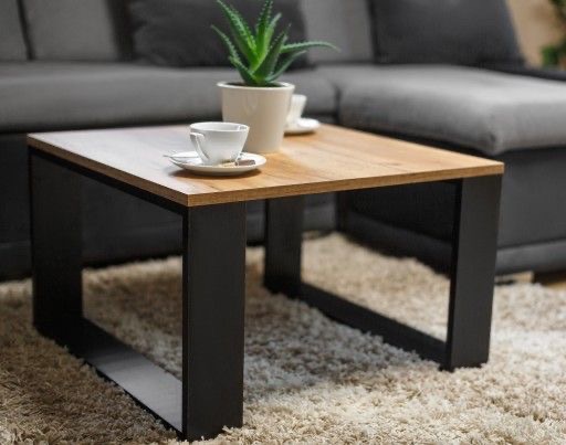 Kwadratowy stolik kawowy z blatem w kolorze jasnego drewna i czarnymi, metalowymi nogami w kształcie litery U, na puszystym dywanie, z filiżanką kawy i rośliną doniczkową na blacie, w tle szara sofa.