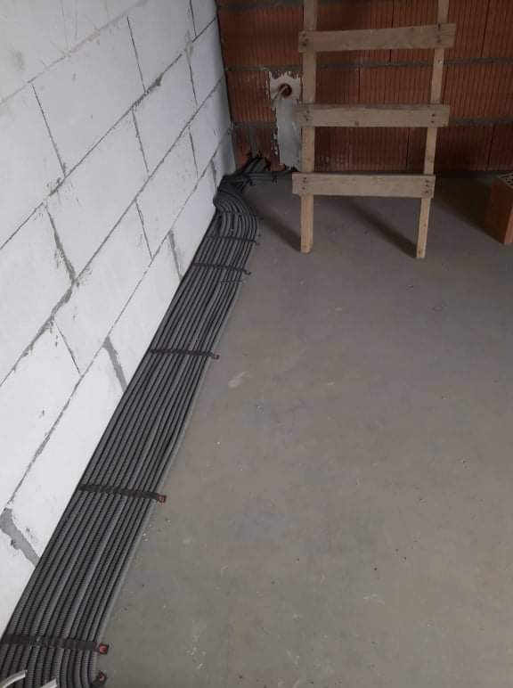 Rzędy szarych rur karbowanych ułożone wzdłuż ściany z białych bloczków, zabezpieczone czarnymi opaskami zaciskowymi, obok drewniana drabina oparta o ścianę z czerwonej cegły w surowym stanie.