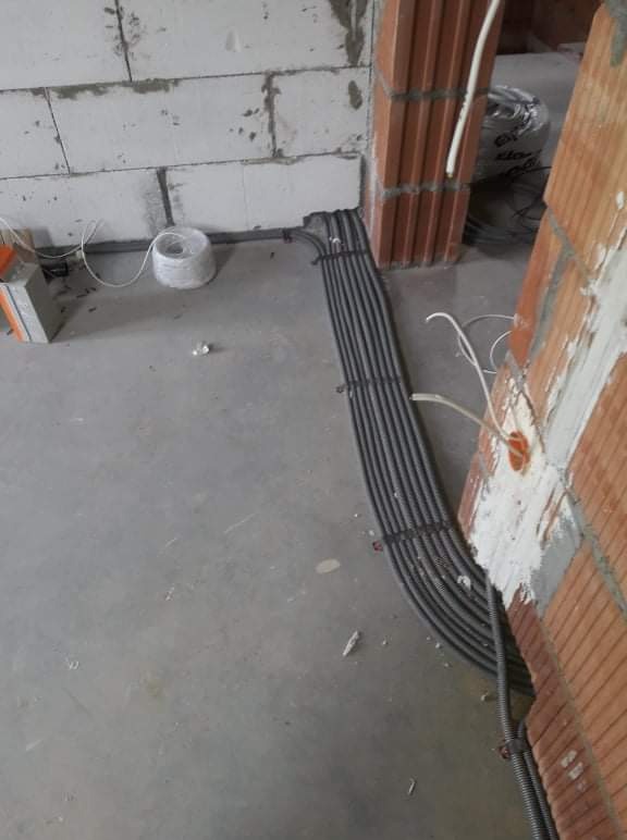 Wiązka szarych peszli z kablami elektrycznymi ułożona wzdłuż ściany z czerwonej cegły dziurawki w surowym stanie, widoczny betonowy podkład i bloczki z betonu komórkowego, instalacja w trakcie...