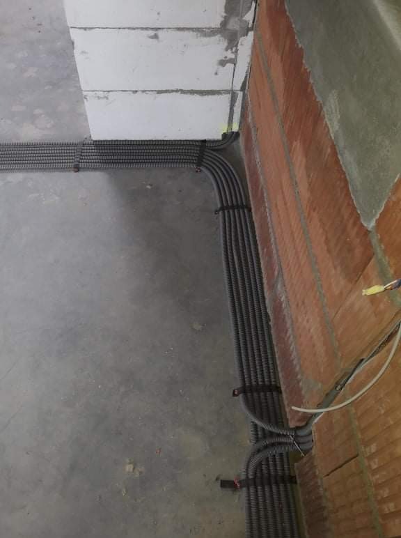 Równe rzędy czarnych, karbowanych peszli elektrycznych ułożone wzdłuż ściany z czerwonej cegły i surowego muru z betonu komórkowego, zabezpieczone czarnymi opaskami zaciskowymi, na szarej...