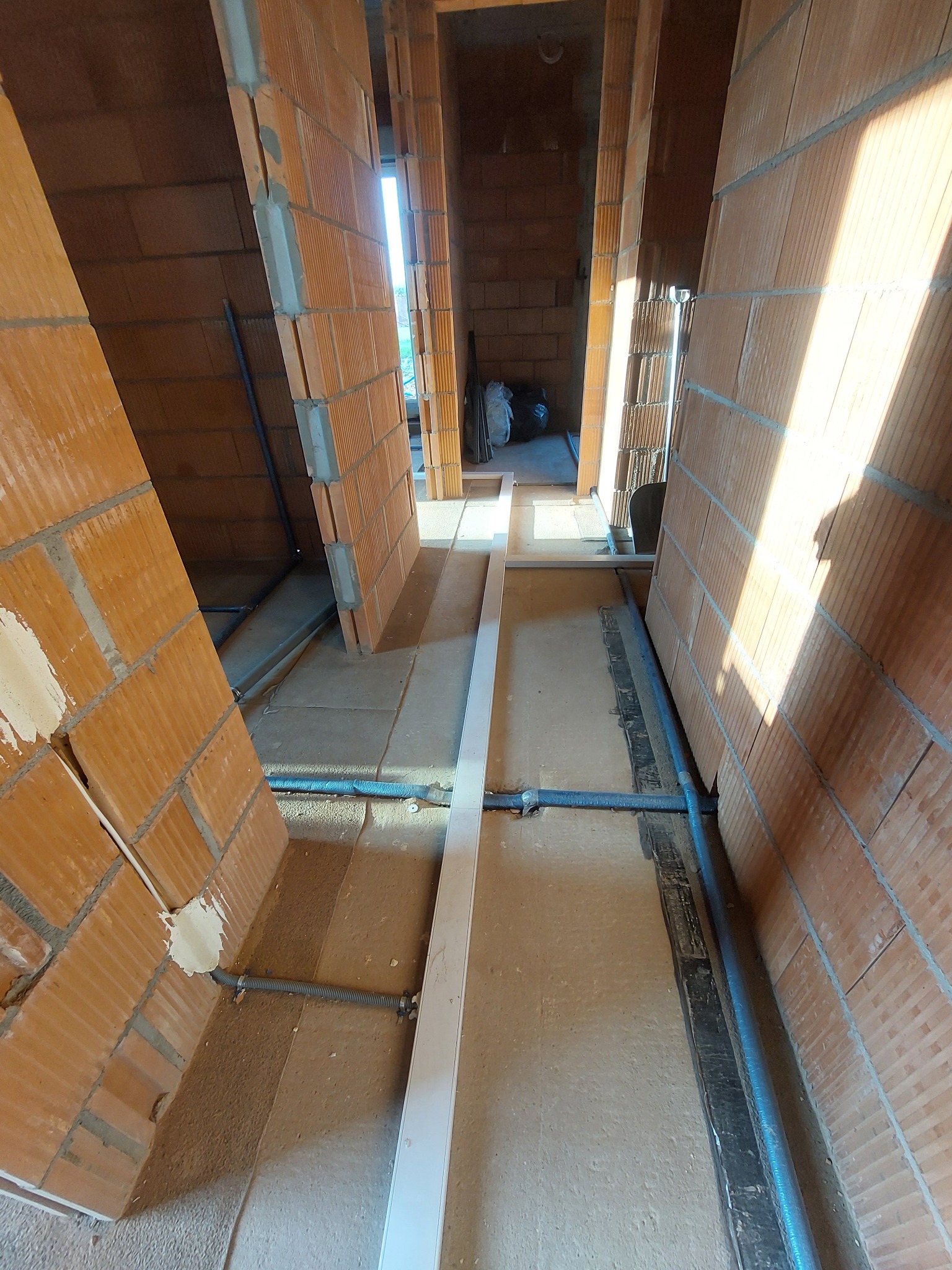 Wewnętrzne ściany z czerwonej cegły dziurawki w trakcie budowy, widoczne szare rury instalacji elektrycznej prowadzone wzdłuż podłogi, biała listwa przypodłogowa na środku kadru, surowe wnętrze...