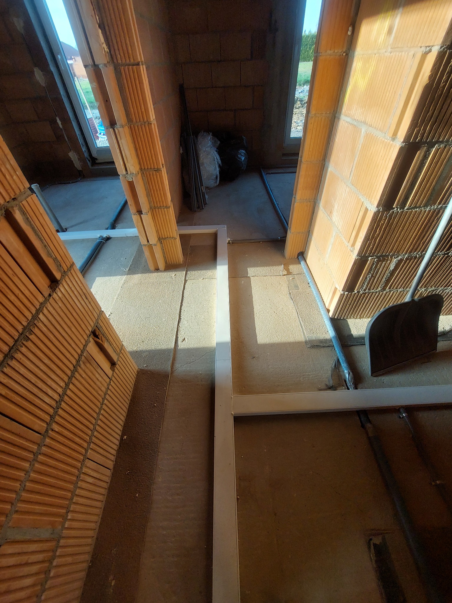 Wnętrze budynku w trakcie budowy: widoczne ceglane ściany, betonowa podłoga, metalowe ramy okienne, instalacja rur elektrycznych i czarny worek na śmieci.