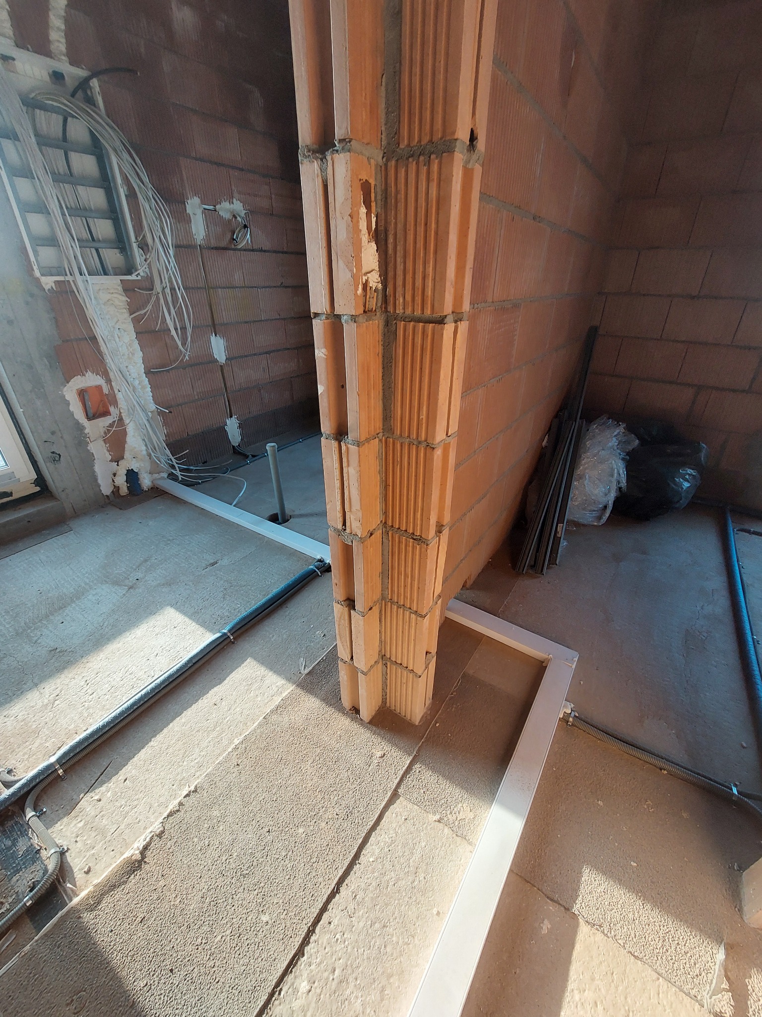 Wnętrze w trakcie budowy z widocznymi przewodami elektrycznymi wychodzącymi ze ściany, rurami ochronnymi na podłodze i fragmentem murowanej ściany z pustaków.