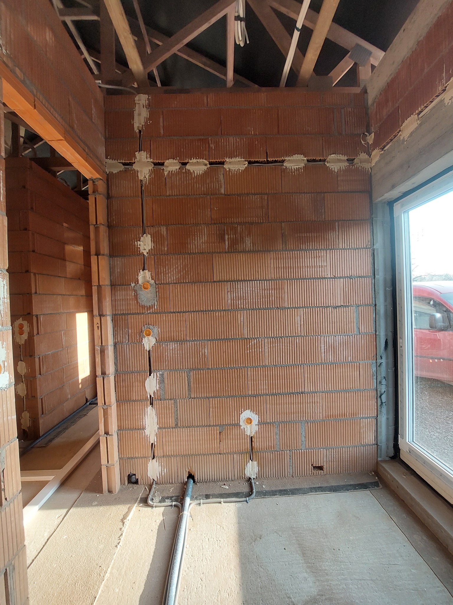 Wnętrze w trakcie budowy z widocznymi przewodami elektrycznymi w ścianach z cegły ceramicznej, zaprawionymi gipsem, oraz rurami instalacyjnymi w podłodze, surowy stan wykończenia.