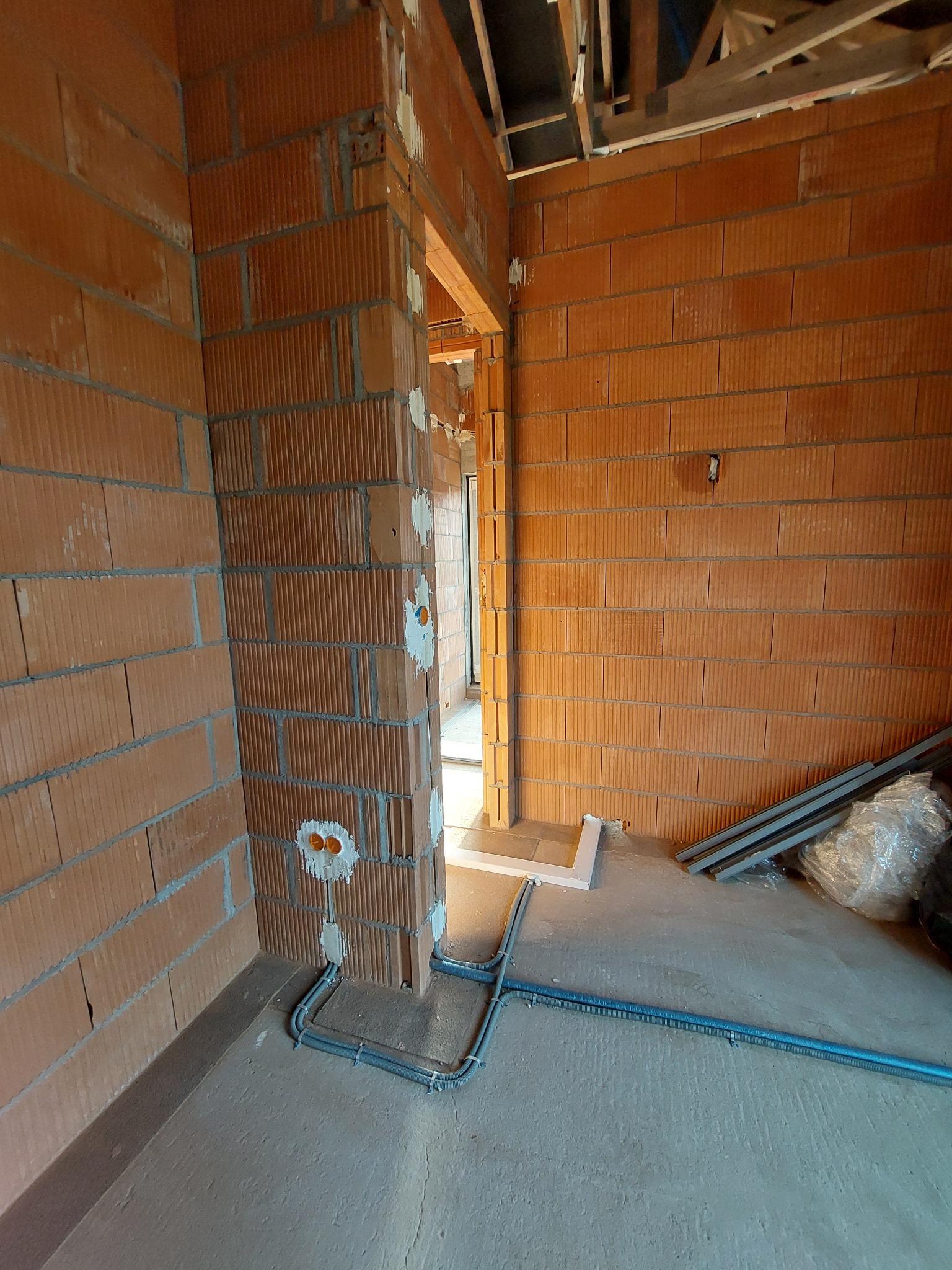 Wnętrze w trakcie budowy z widocznymi przewodami elektrycznymi w szarych rurkach mocowanych do ściany z czerwonej cegły, puszki elektryczne częściowo zamurowane.