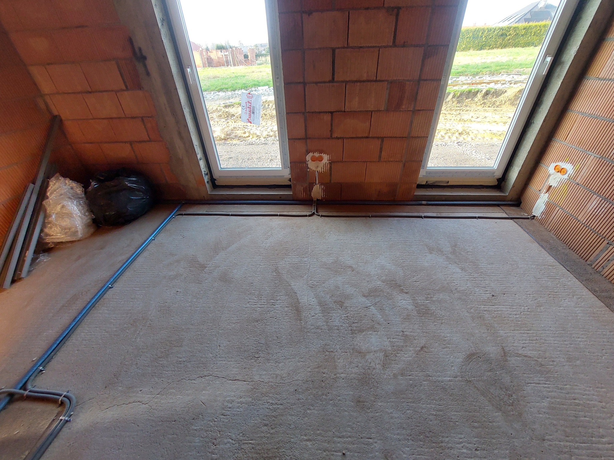 Surowy stan poddasza z dwoma oknami dachowymi, widoczne kable elektryczne poprowadzone wzdłuż podłogi i ścian z pustaków ceramicznych. W tle widok z okna na budowę i zieleń.
