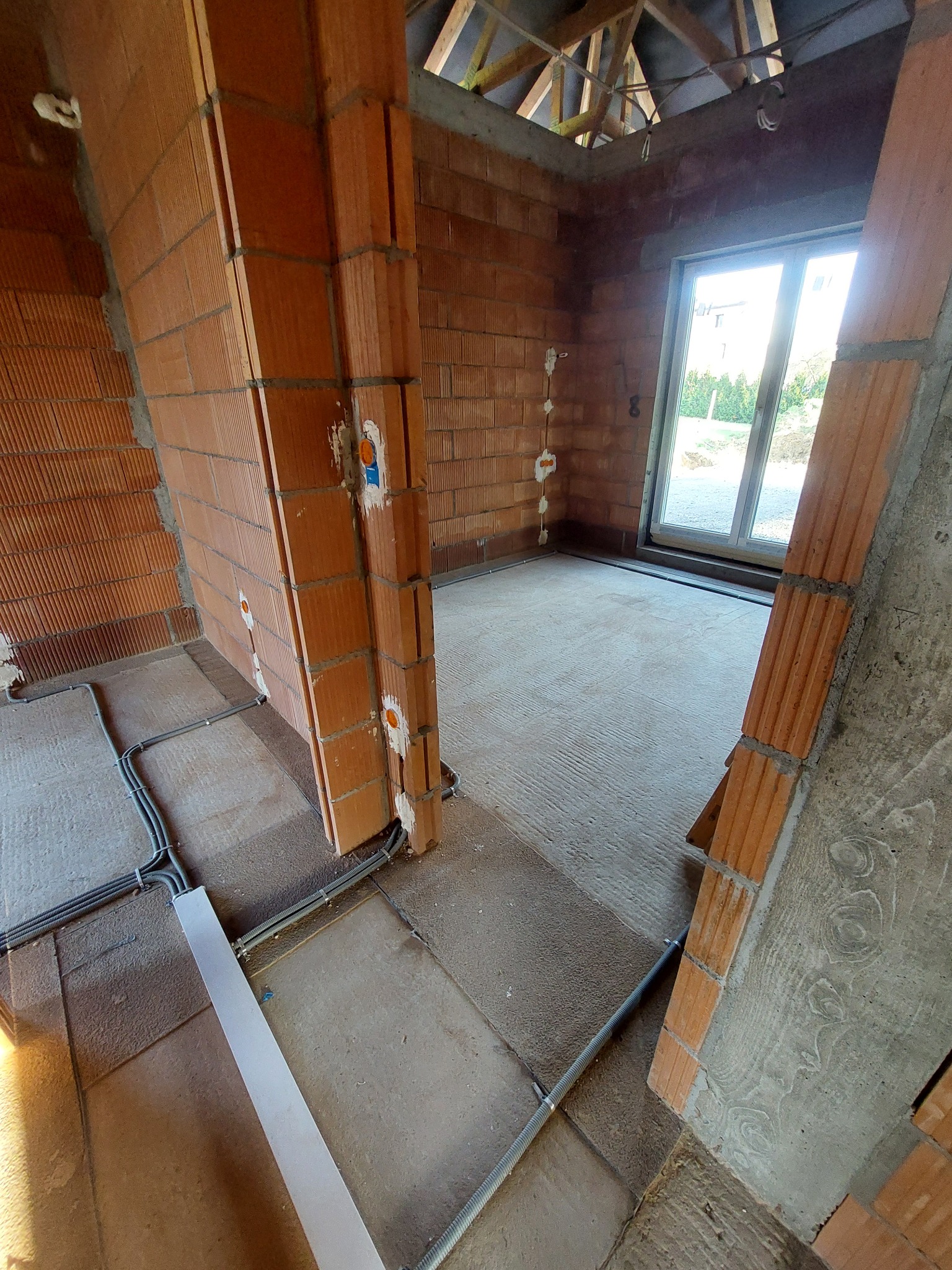 Wnętrze domu w trakcie budowy z widocznymi przewodami elektrycznymi w peszlach ułożonymi na podłodze, ceglane ściany w surowym stanie z puszkami instalacyjnymi.