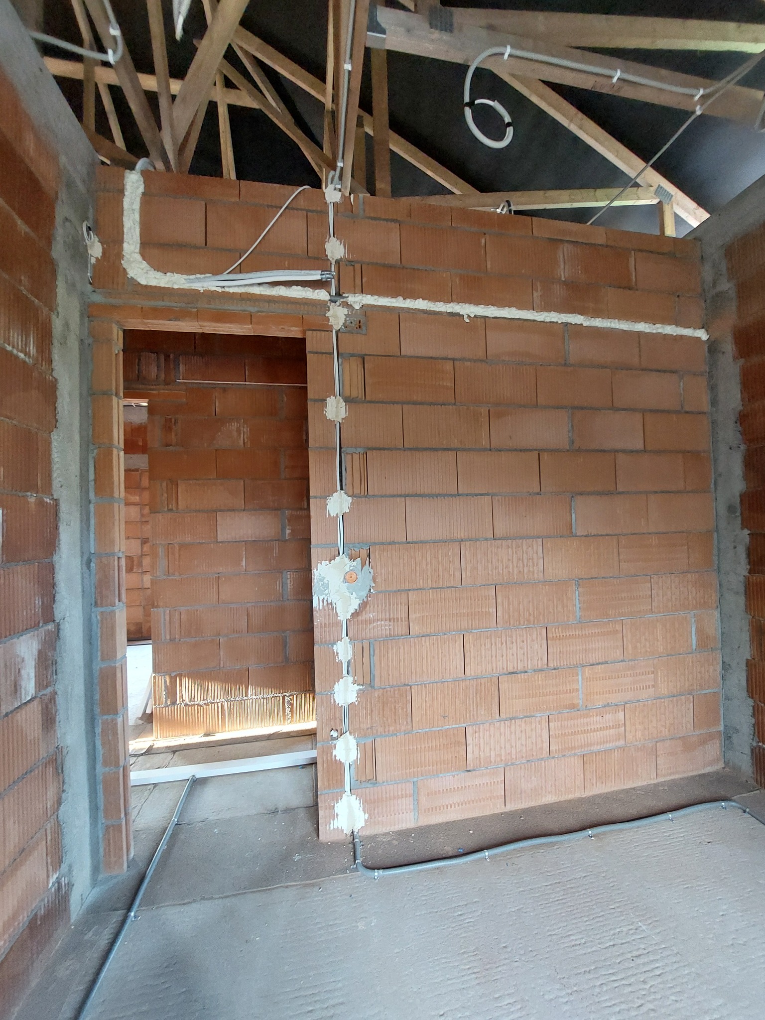 Wnętrze w trakcie budowy, widoczne przewody elektryczne zabezpieczone pianką montażową na ścianach z czerwonej cegły, drewniana konstrukcja dachu w tle.