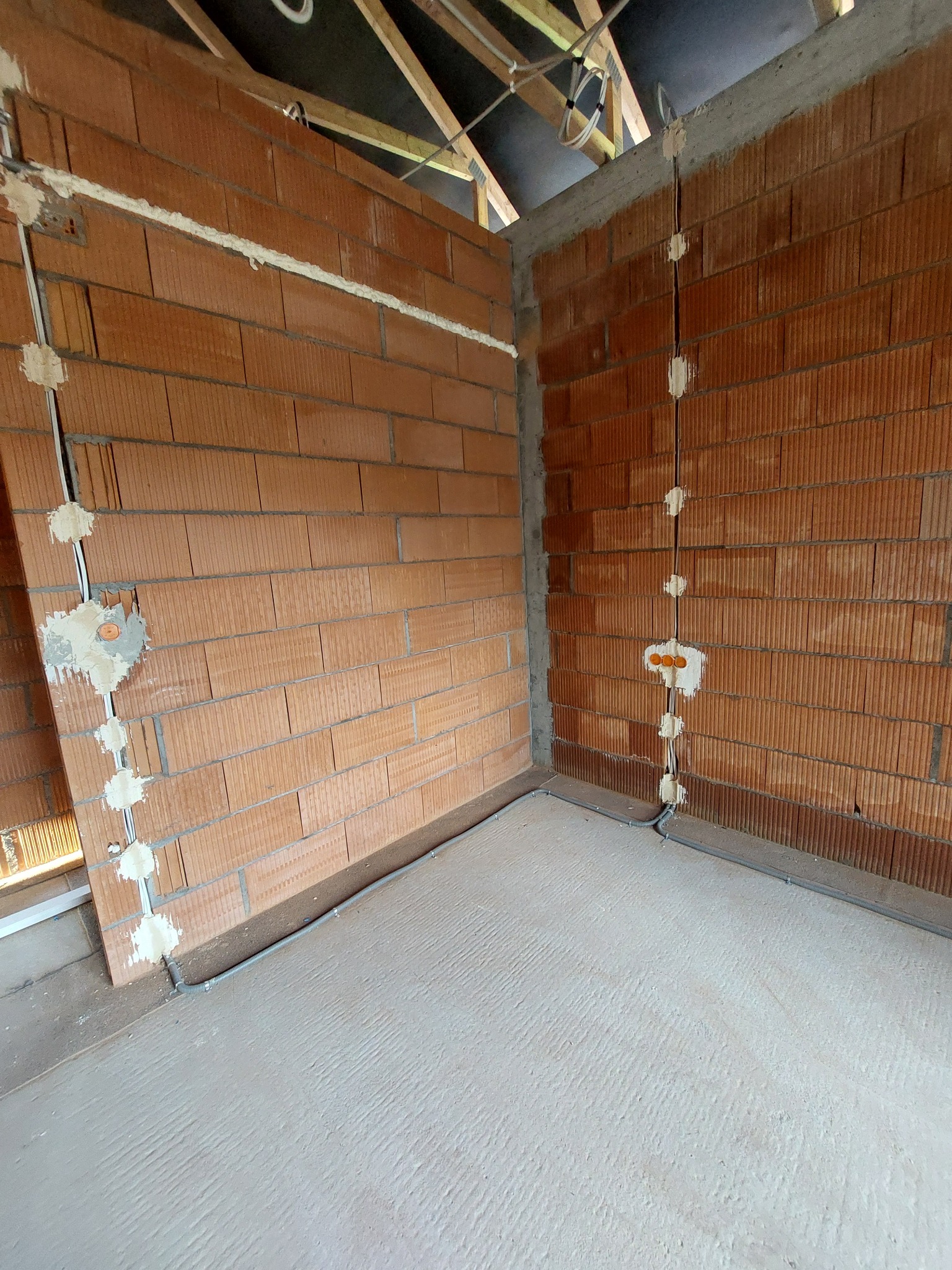 Wnętrze w stanie surowym: murowane ściany z czerwonej cegły, wstępnie poprowadzone przewody elektryczne w bruzdach zabezpieczone gipsem, widoczne elementy konstrukcji dachu.