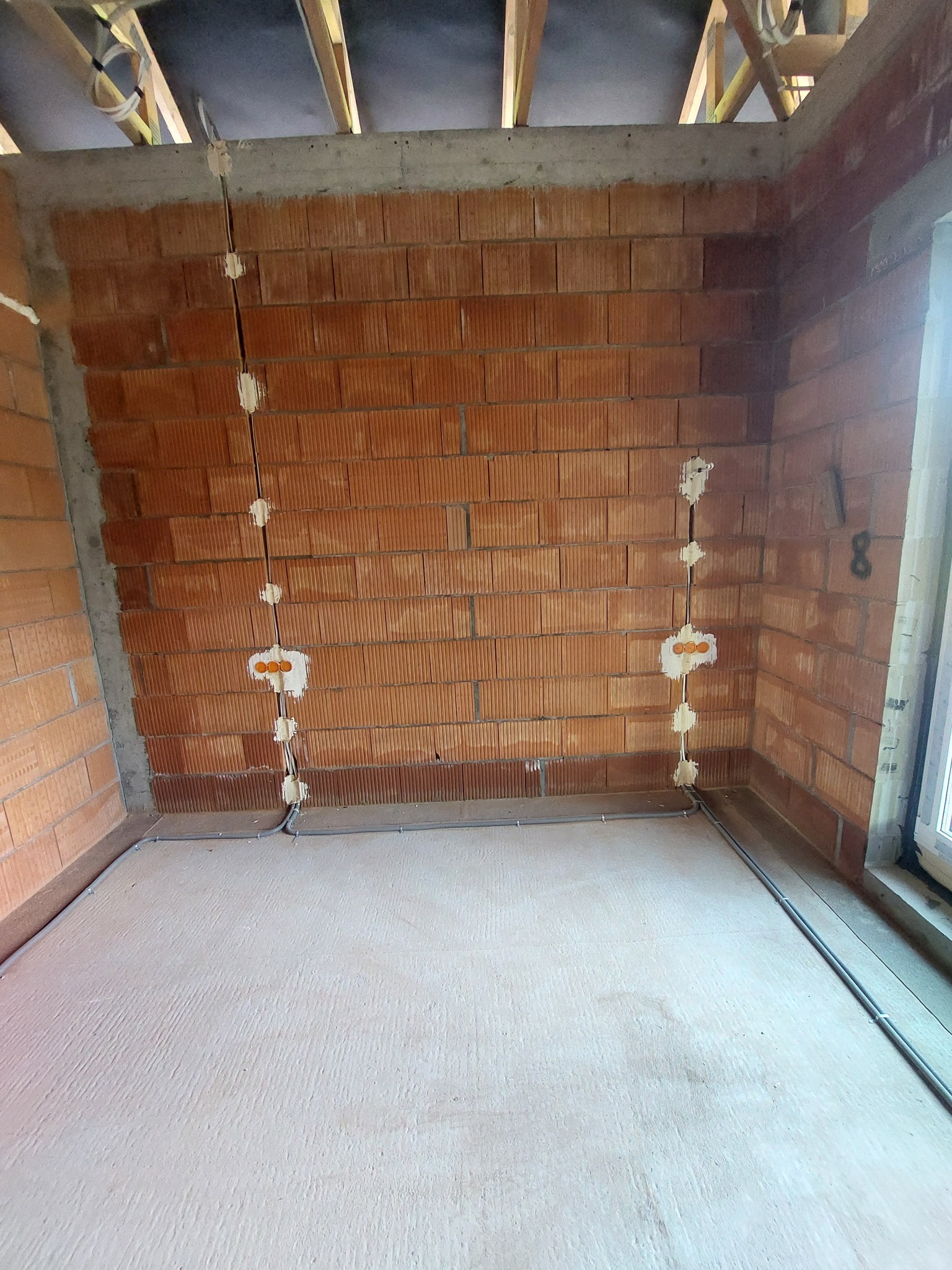 Surowy mur z cegły z przewodami elektrycznymi w peszlach, częściowo zamaskowanymi pianką montażową, widoczne puszki instalacyjne z pomarańczowymi wkładami, szare rurki peszel prowadzone wzdłuż...