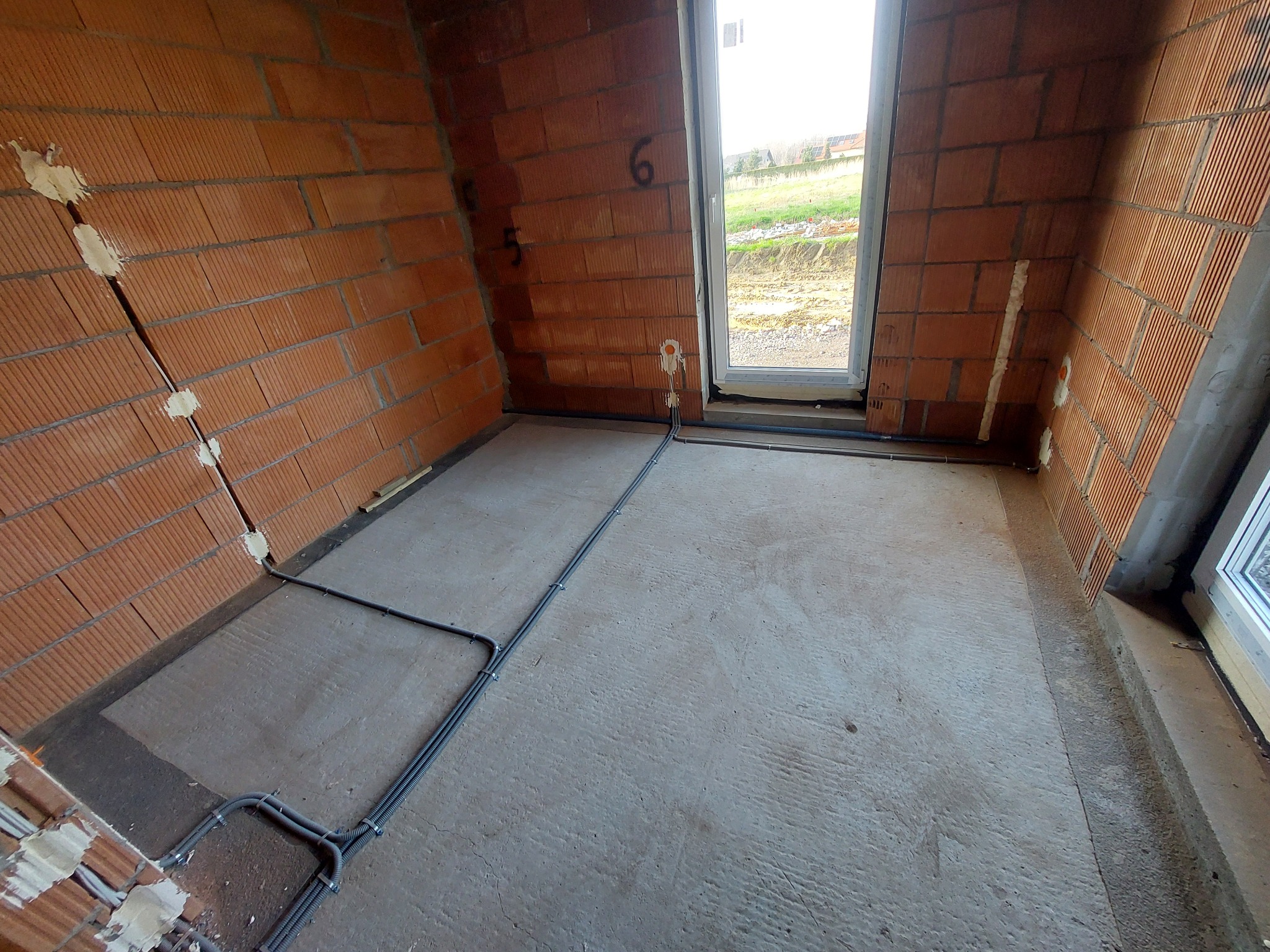 Wnętrze w stanie surowym z widoczną instalacją elektryczną w peszlach, prowadzoną po podłodze i ścianach z cegły, z widocznymi puszkami instalacyjnymi w ścianach i oknem w tle.