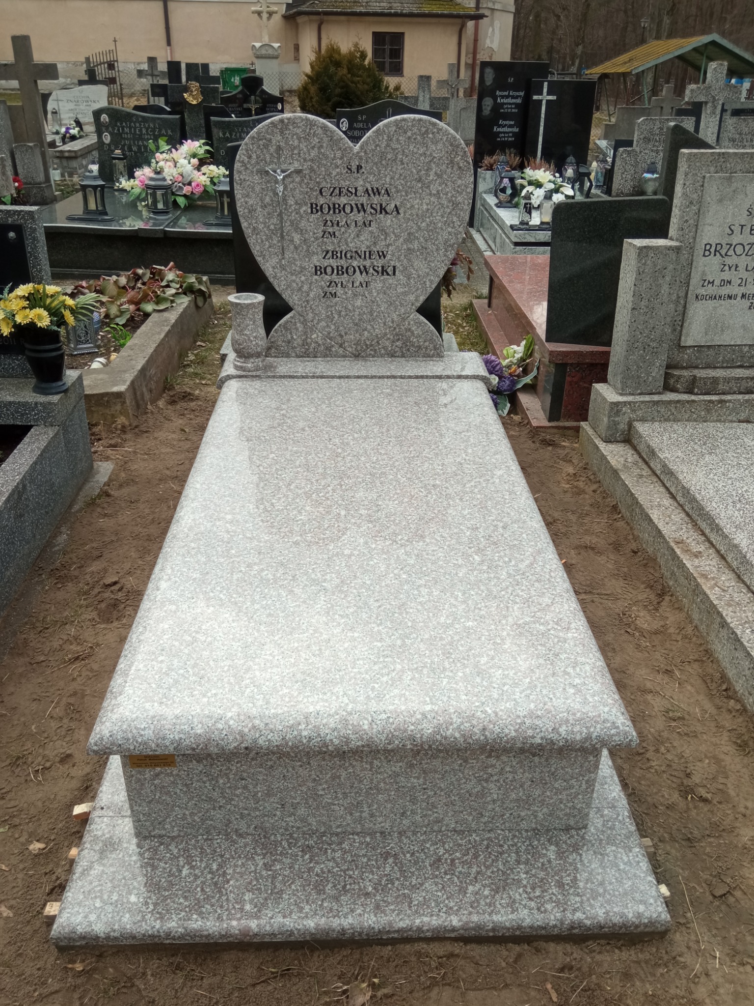 Jasnoszary granitowy pomnik z płytą i sercem na cmentarzu, z wygrawerowanymi imionami i nazwiskami, otoczony ziemią i innymi nagrobkami.