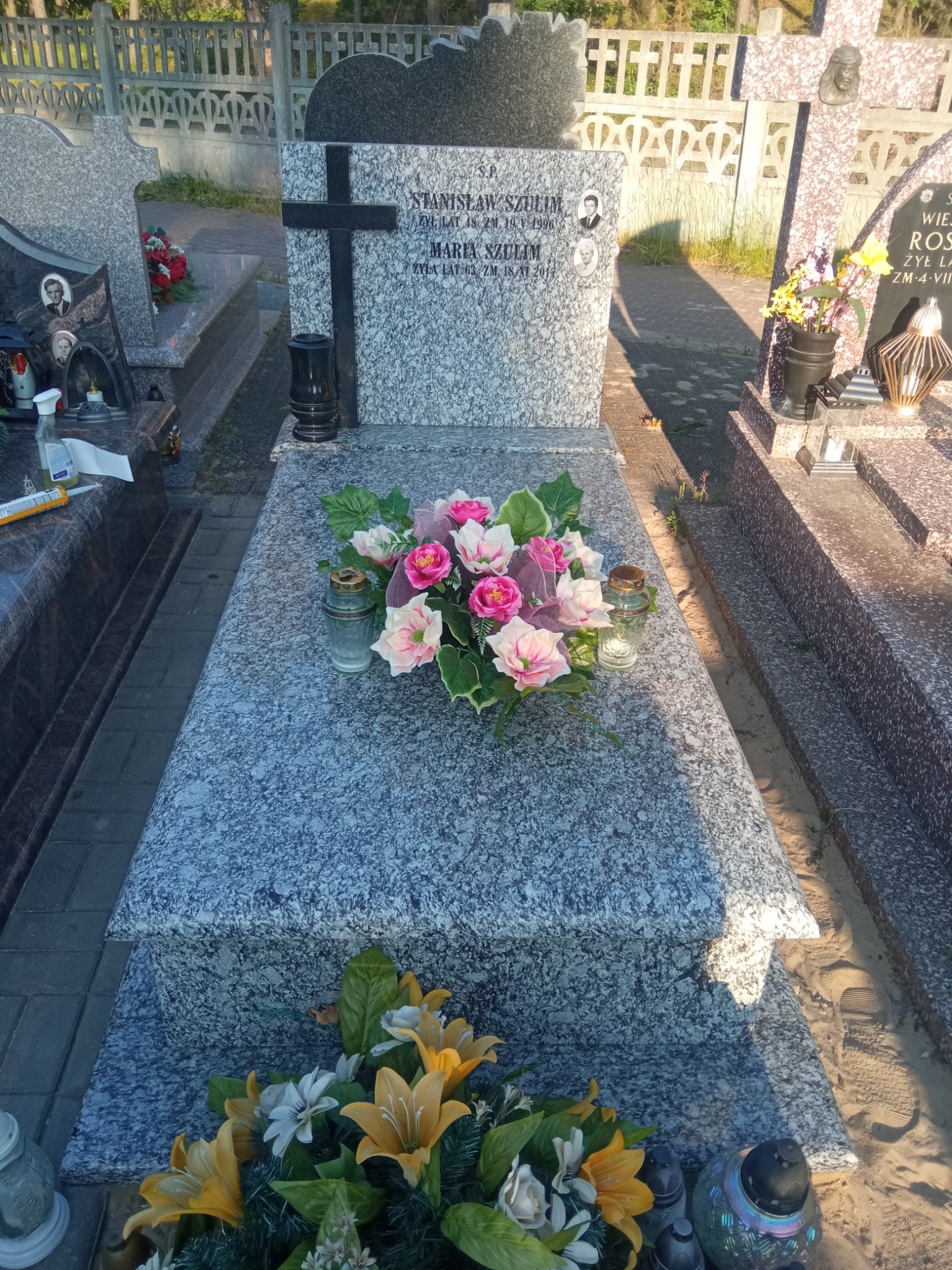 Granitowy pomnik z ciemnym krzyżem i portretami Stanisława i Marii Szulim, ozdobiony bukietem różowych i białych kwiatów oraz lampionami, na tle innych nagrobków.