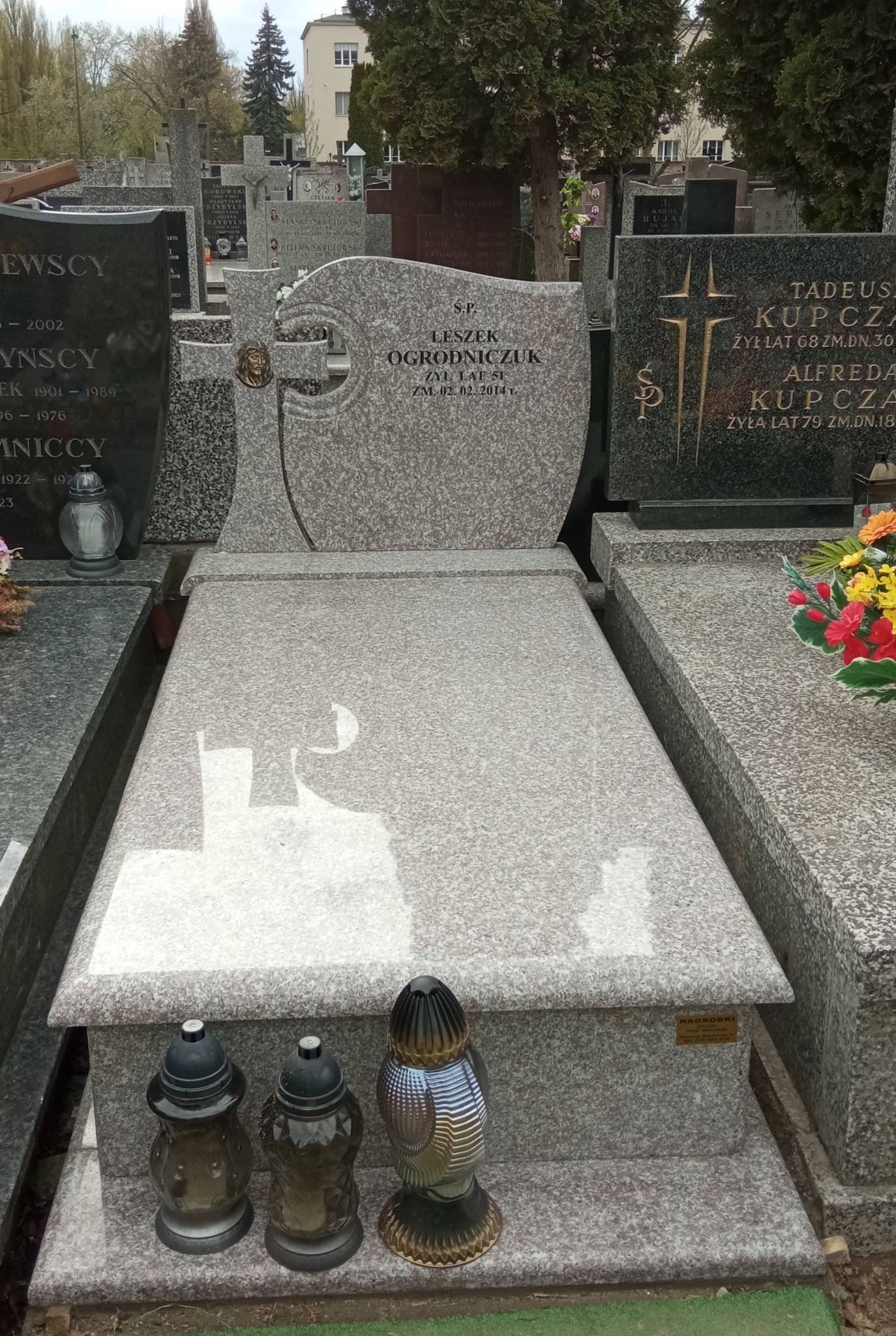 Jasnoszary, granitowy pomnik z wygrawerowanym imieniem i nazwiskiem Leszek Ogrodniczuk oraz datą śmierci 02.02.2014, widoczne trzy znicze na dole pomnika, cmentarne otoczenie w tle.