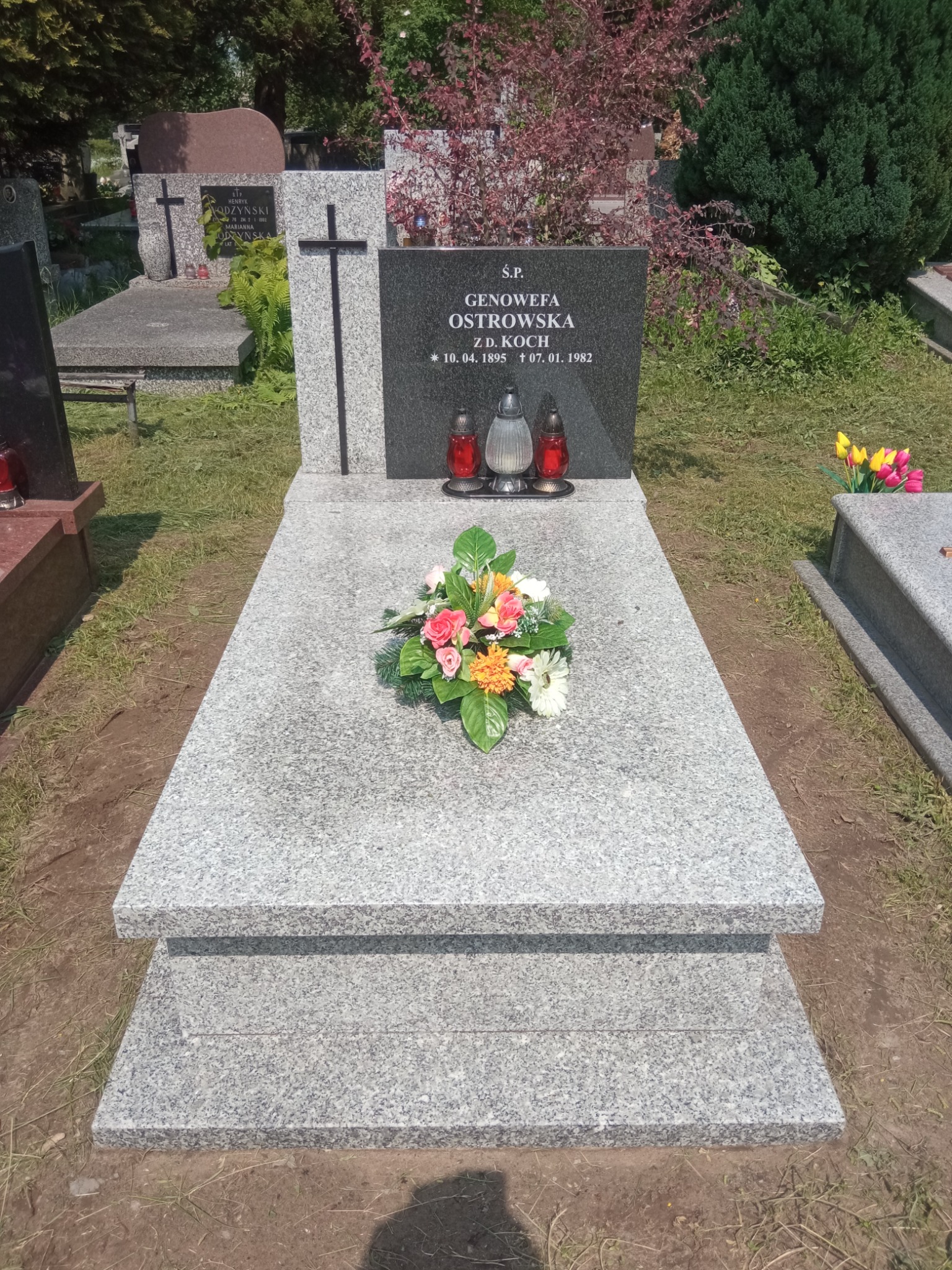 Granitowy pomnik z dwoma zniczami i bukietem kwiatów na cmentarzu, w tle inne groby i zieleń.