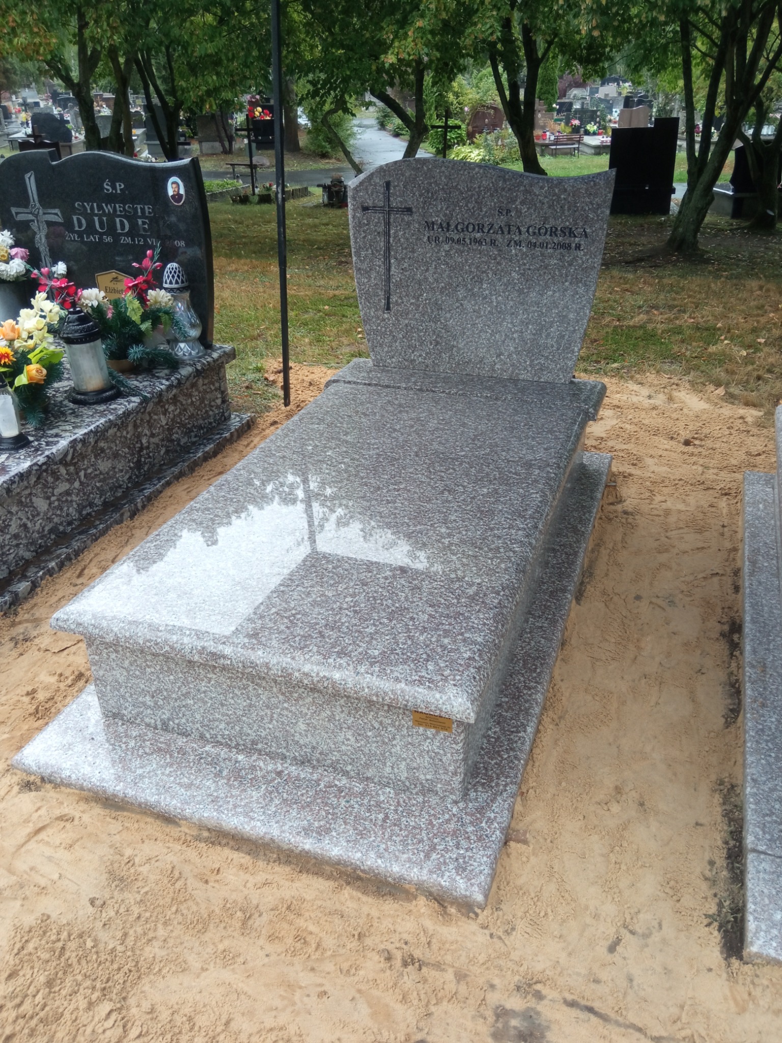 Nowo postawiony, trzyczęściowy, szary pomnik z granitu na cmentarzu, z inskrypcją i krzyżem, otoczony piaskiem.