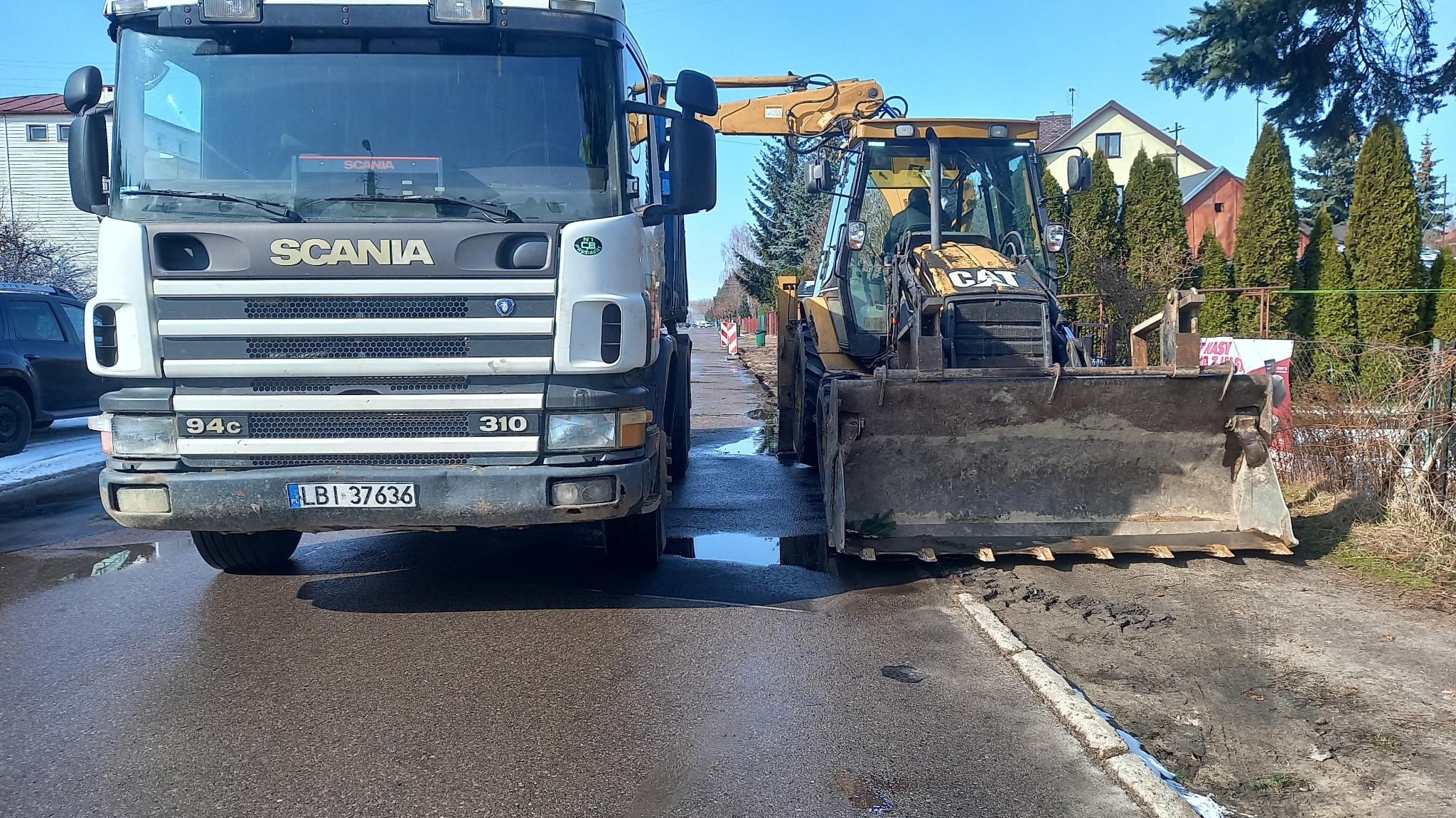 Ciężarówka Scania i koparko-ładowarka Caterpillar stojące na mokrej, asfaltowej drodze w dzielnicy mieszkalnej, prawdopodobnie po deszczu.