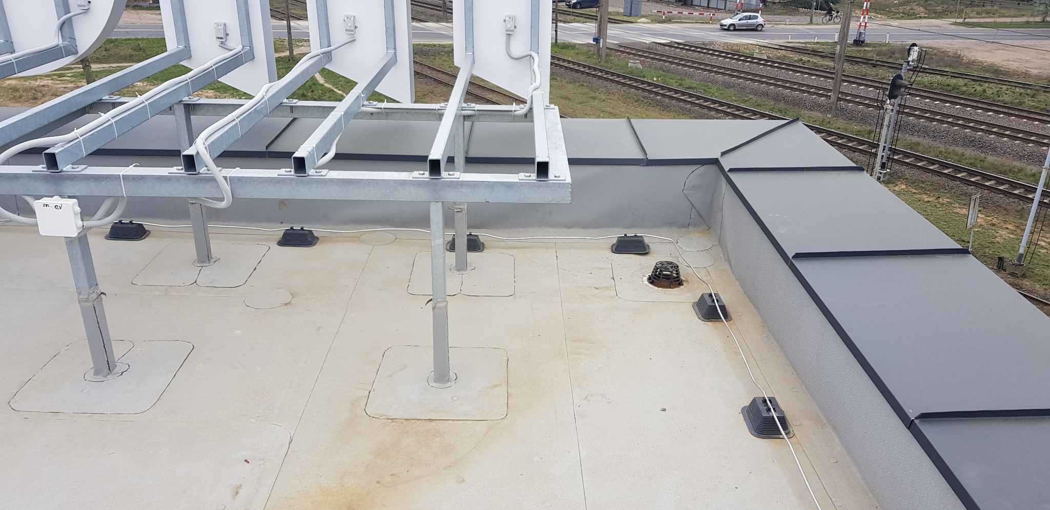 Płaski dach pokryty szarym materiałem z widoczną konstrukcją wsporczą pod panele słoneczne oraz elementami wentylacyjnymi, w tle tory kolejowe.