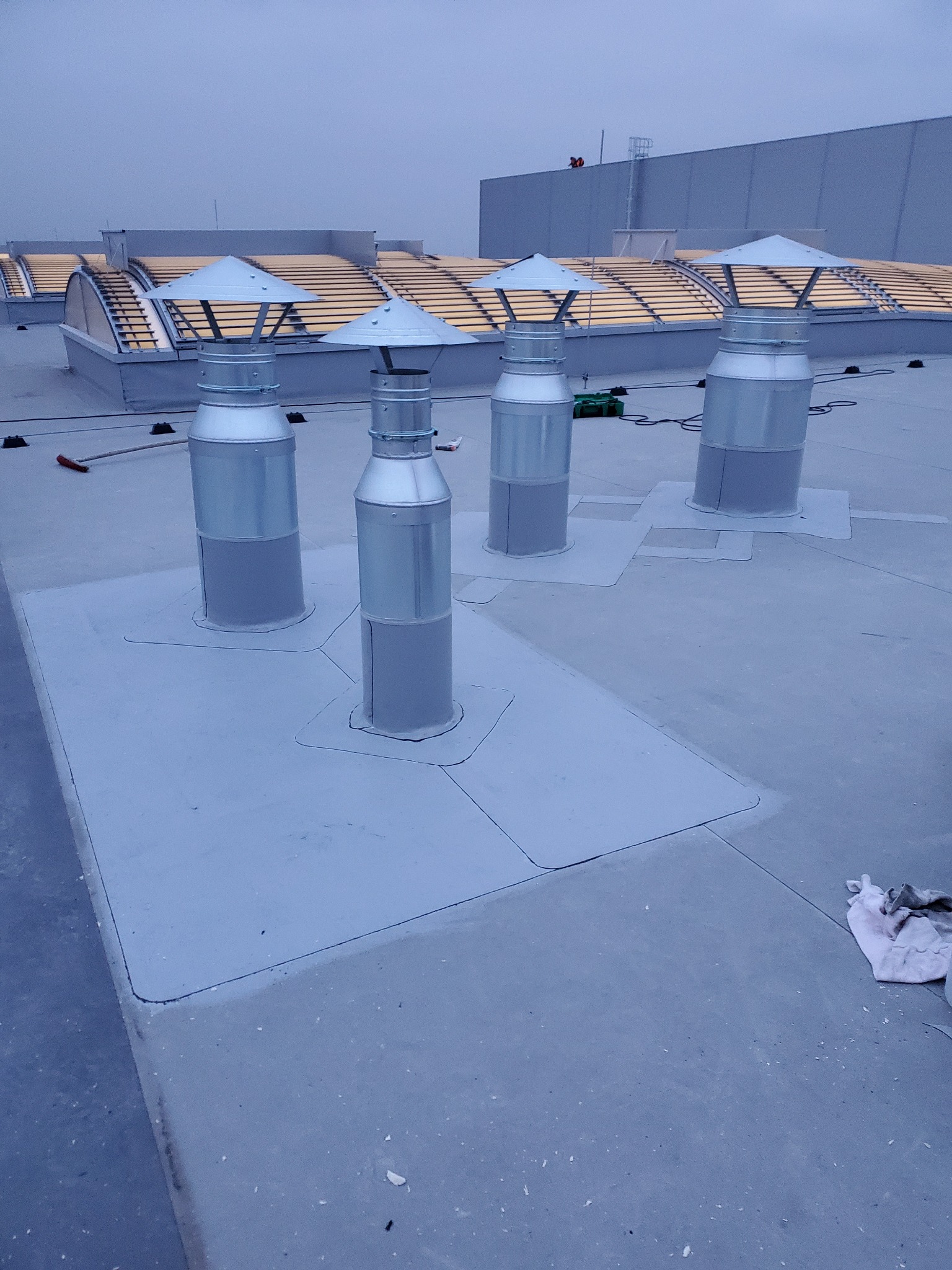 Płaski dach przemysłowy z czterema metalowymi kominami wentylacyjnymi, świeżo uszczelniony szarą powłoką, z widocznymi świetlikami w tle i narzędziami pracy.