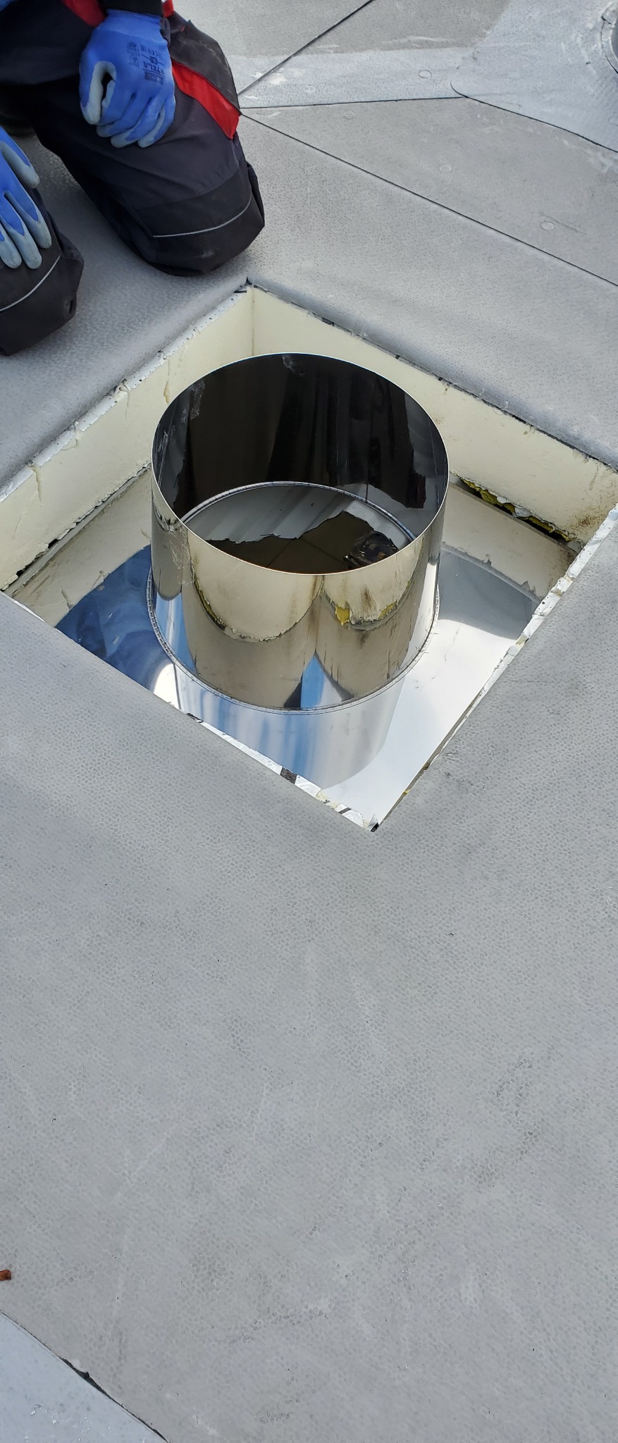 Montaż metalowej obróbki komina na płaskim dachu z widocznymi warstwami izolacji i pracownikiem w rękawicach ochronnych w tle.