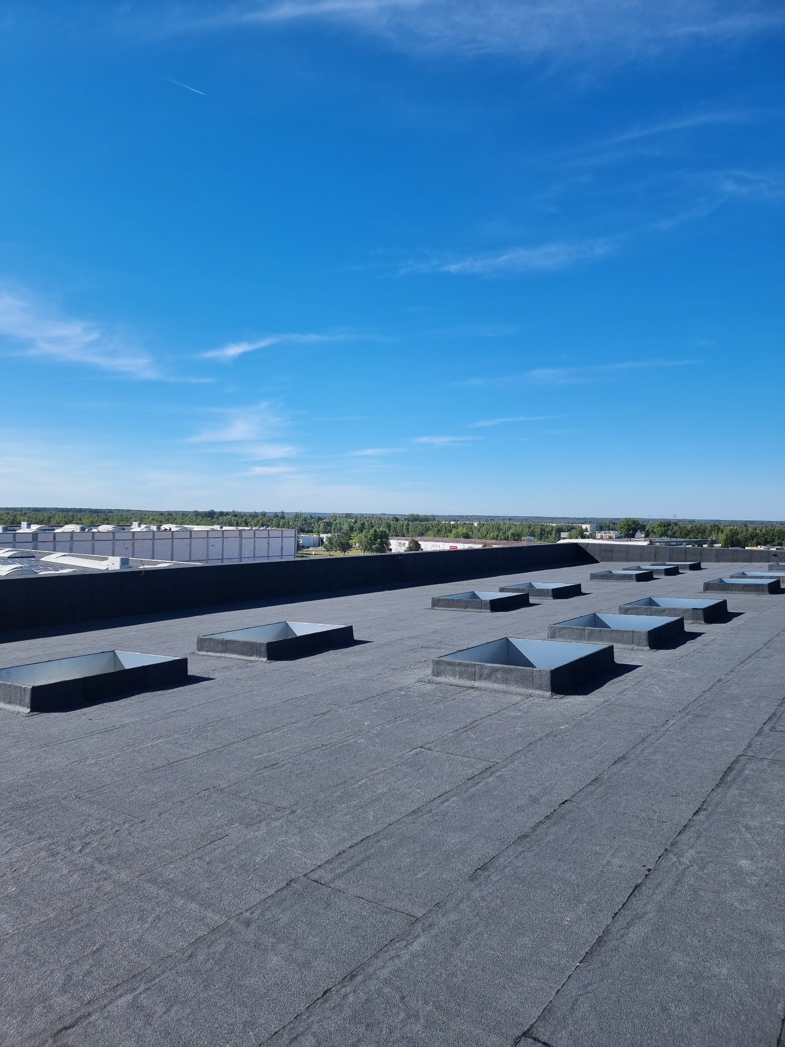 Bitumiczna powierzchnia dachu przemysłowego z świetlikami w kształcie kwadratów, widok na odległy horyzont i błękitne niebo z delikatnymi chmurami.