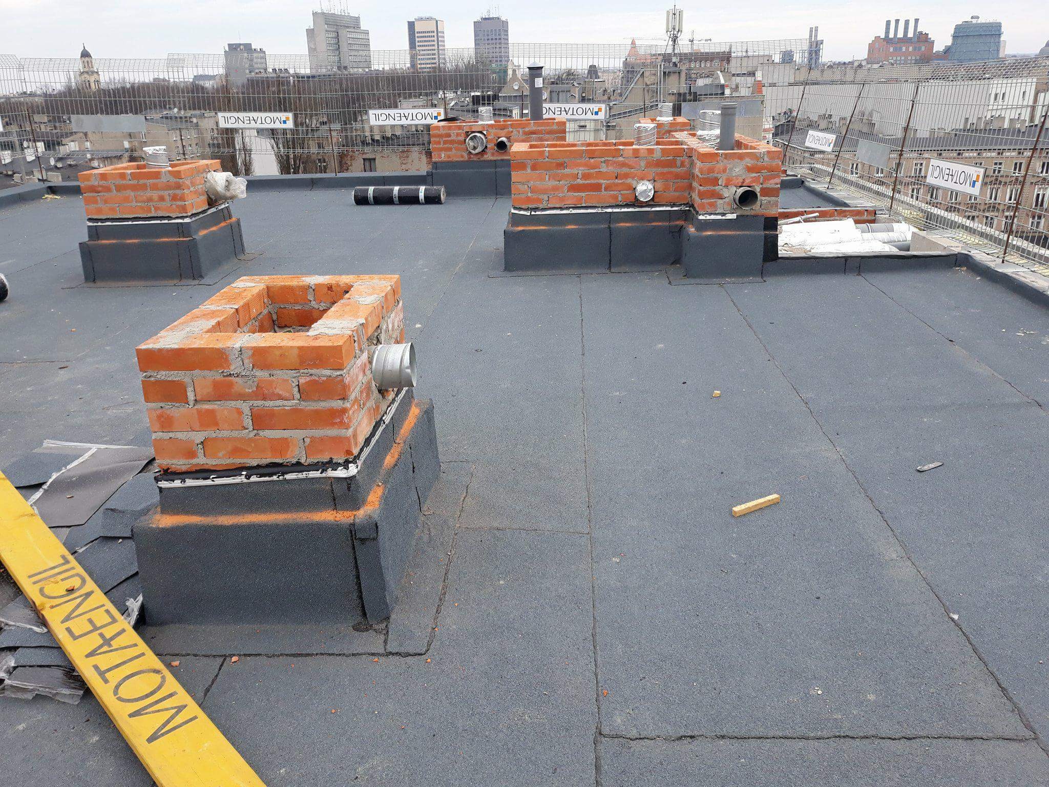 Widok z góry na płaski dach pokryty papą z wystającymi ceglanymi kominami w trakcie budowy, z widoczną miejską panoramą w tle.