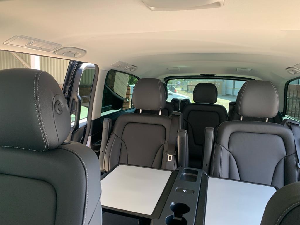 Wnętrze luksusowego vana z czarnymi skórzanymi fotelami, rozkładanymi stolikami i uchwytami na napoje, widok z tyłu pojazdu.