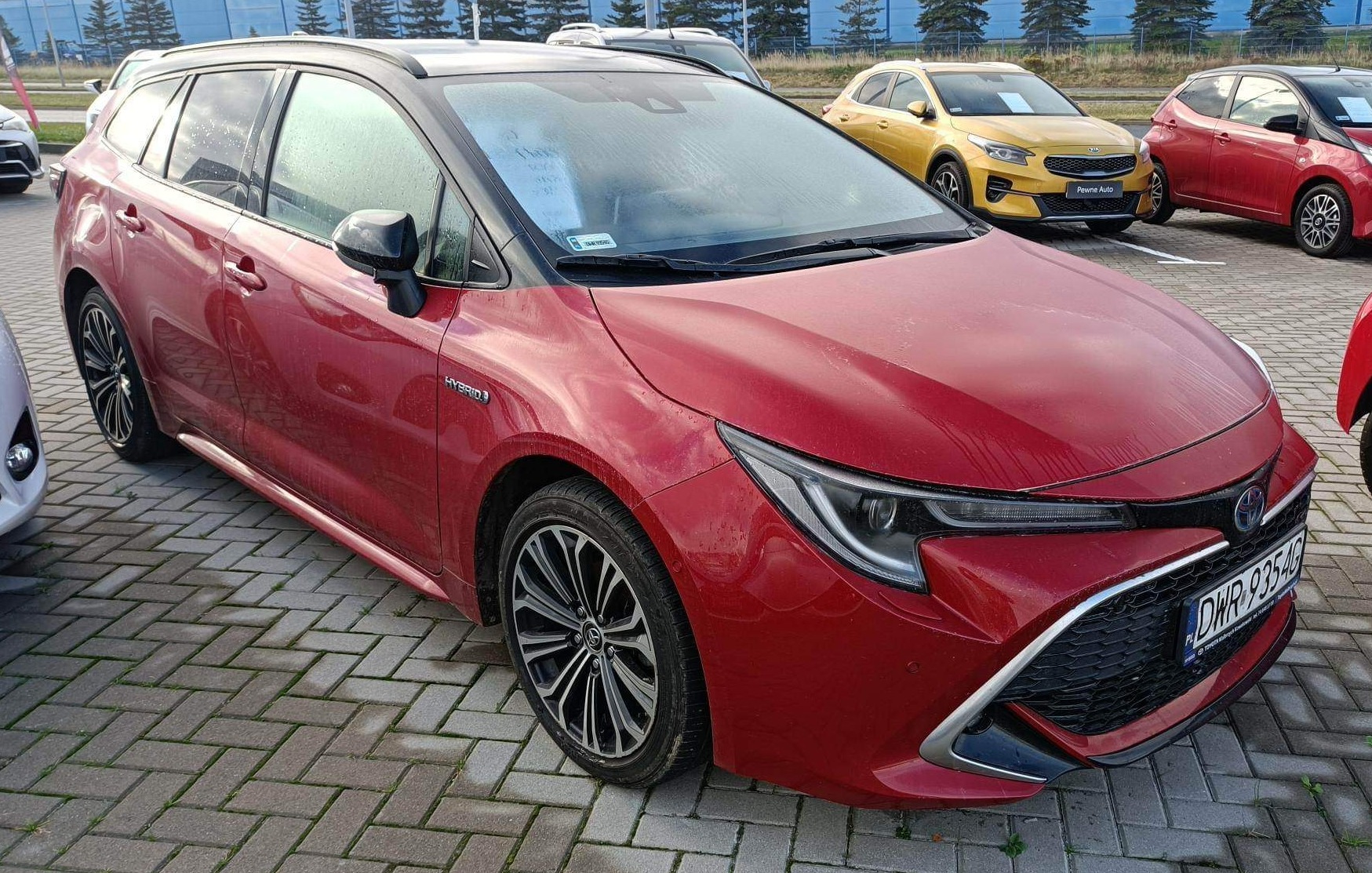 Czerwony samochód typu kombi z napędem hybrydowym, zaparkowany na brukowanym parkingu, widoczny z przodu i boku, z innymi autami w tle.