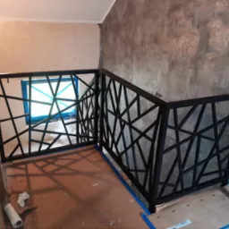 Metalowa balustrada schodowa o geometrycznym wzorze, czarna, zamontowana w surowym wnętrzu z szarą, teksturowaną ścianą i zabezpieczonymi folią oknami, widoczne ślady prac wykończeniowych.