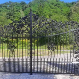 Elegancka, czarna, kuta brama wjazdowa z motywem winorośli, zamontowana między dwoma kamiennymi słupkami z dekoracyjnymi zwieńczeniami, z widocznym mechanizmem automatycznego otwierania.