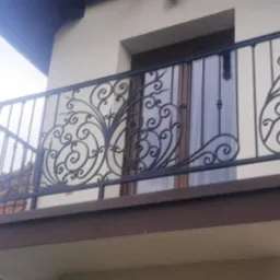 Kuta balustrada balkonowa z dekoracyjnymi elementami w kształcie roślinnych ornamentów na tle okna z zasłoną.