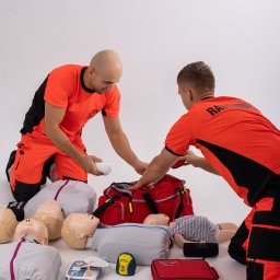 Med4Life Andrzej Olejarz - Dwóch ratowników w pomarańczowych uniformach przygotowuje torbę medyczną na białym tle, otoczoną manekinami do ćwiczeń resuscytacji, defibrylatorem i akcesoriami.