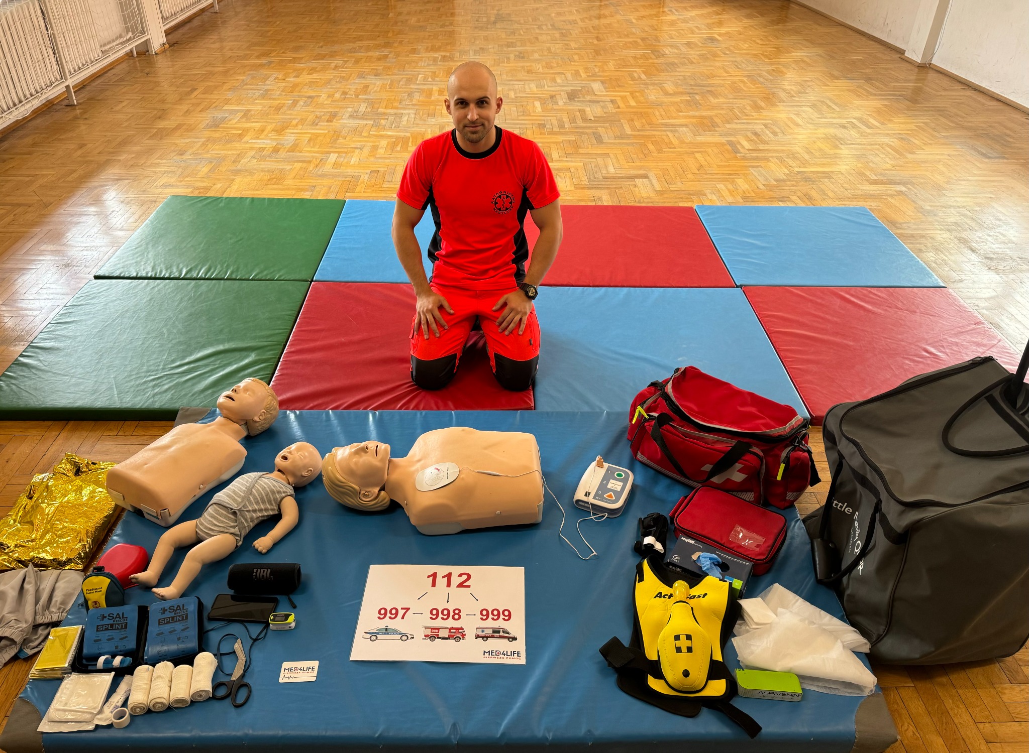 Instruktor klęczy na kolorowych matach, prezentując manekiny niemowląt i dorosłych, sprzęt medyczny, w tym defibrylator i torby ratunkowe, oraz kartę z numerami alarmowymi 112, 997, 998 i 999.