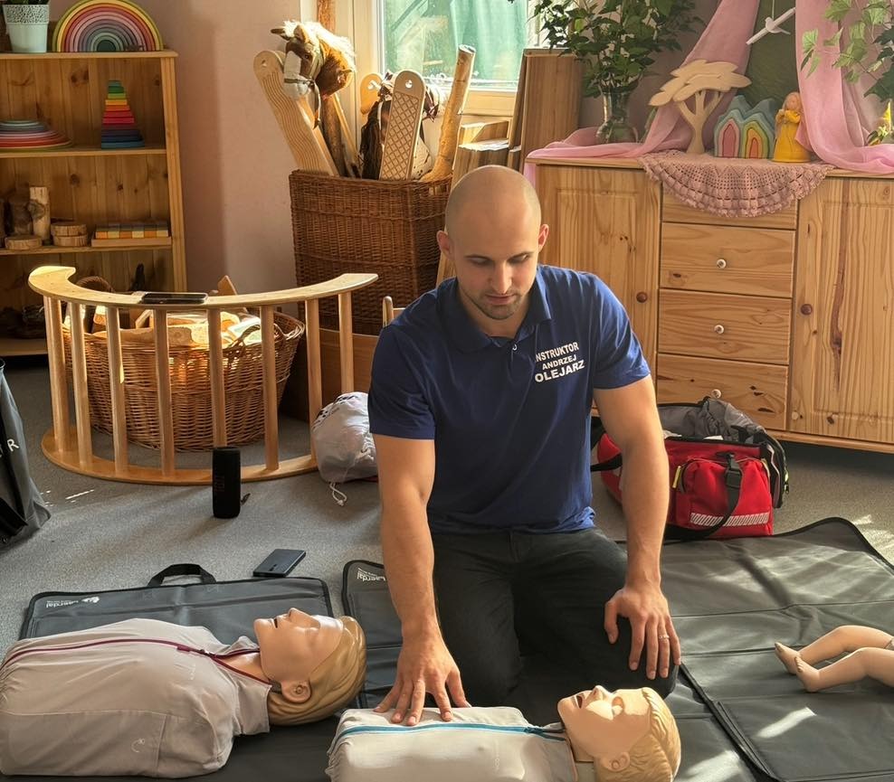 Instruktor w niebieskiej koszulce z napisem 'INSTRUKTOR ANDRZEJ OLEJARZ' demonstruje resuscytację krążeniowo-oddechową na manekinach na macie, w tle drewniane meble i zabawki.