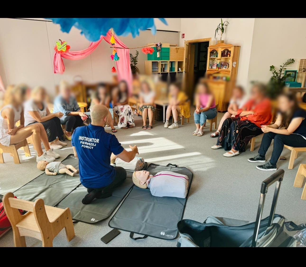 Sala z grupą osób siedzących na krzesłach wokół instruktora demonstrującego resuscytację na manekinach, widoczne też walizki i dekoracje w tle.