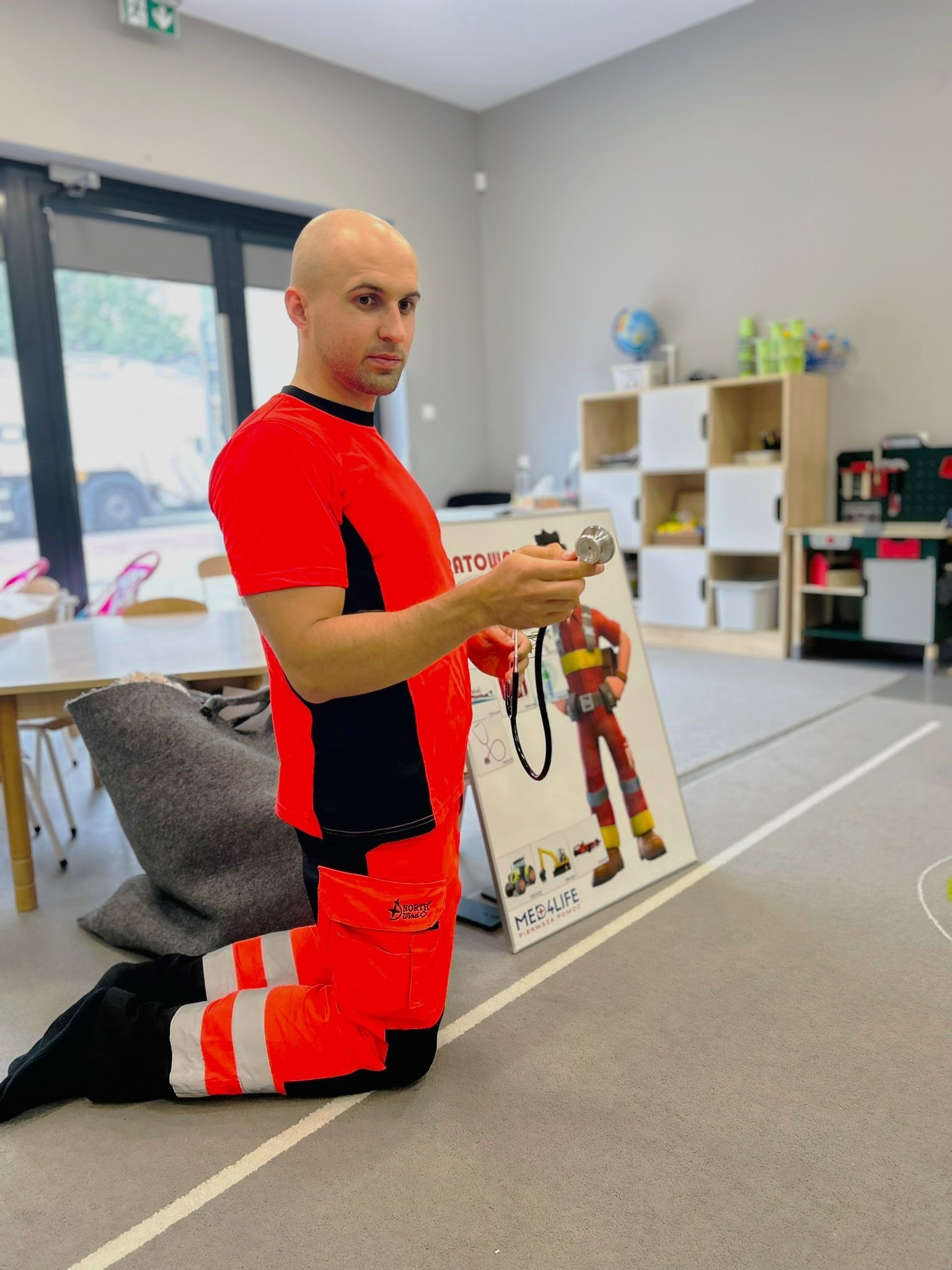 Instruktor w stroju ratownika medycznego na kolanach trzyma stetoskop, obok plansza edukacyjna z postacią ratownika i pojazdami budowlanymi.