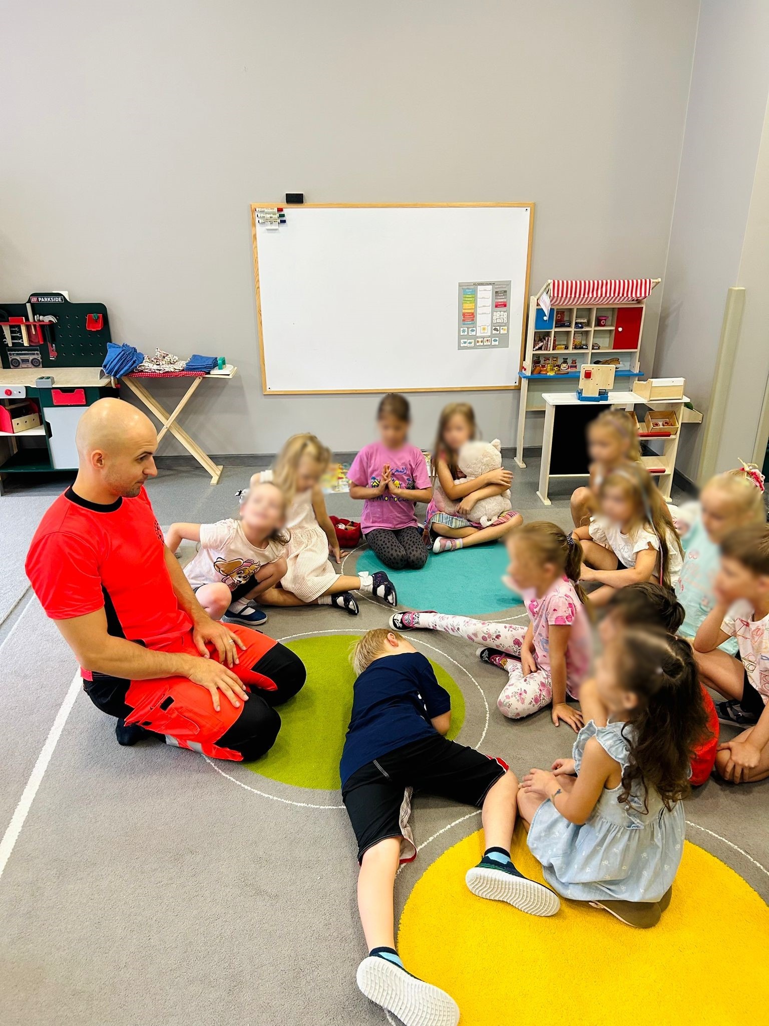 Instruktor w czerwonym stroju demonstruje pozycję bezpieczną uczniowi podczas kursu pierwszej pomocy dla dzieci, siedzących na kolorowym dywanie w sali.