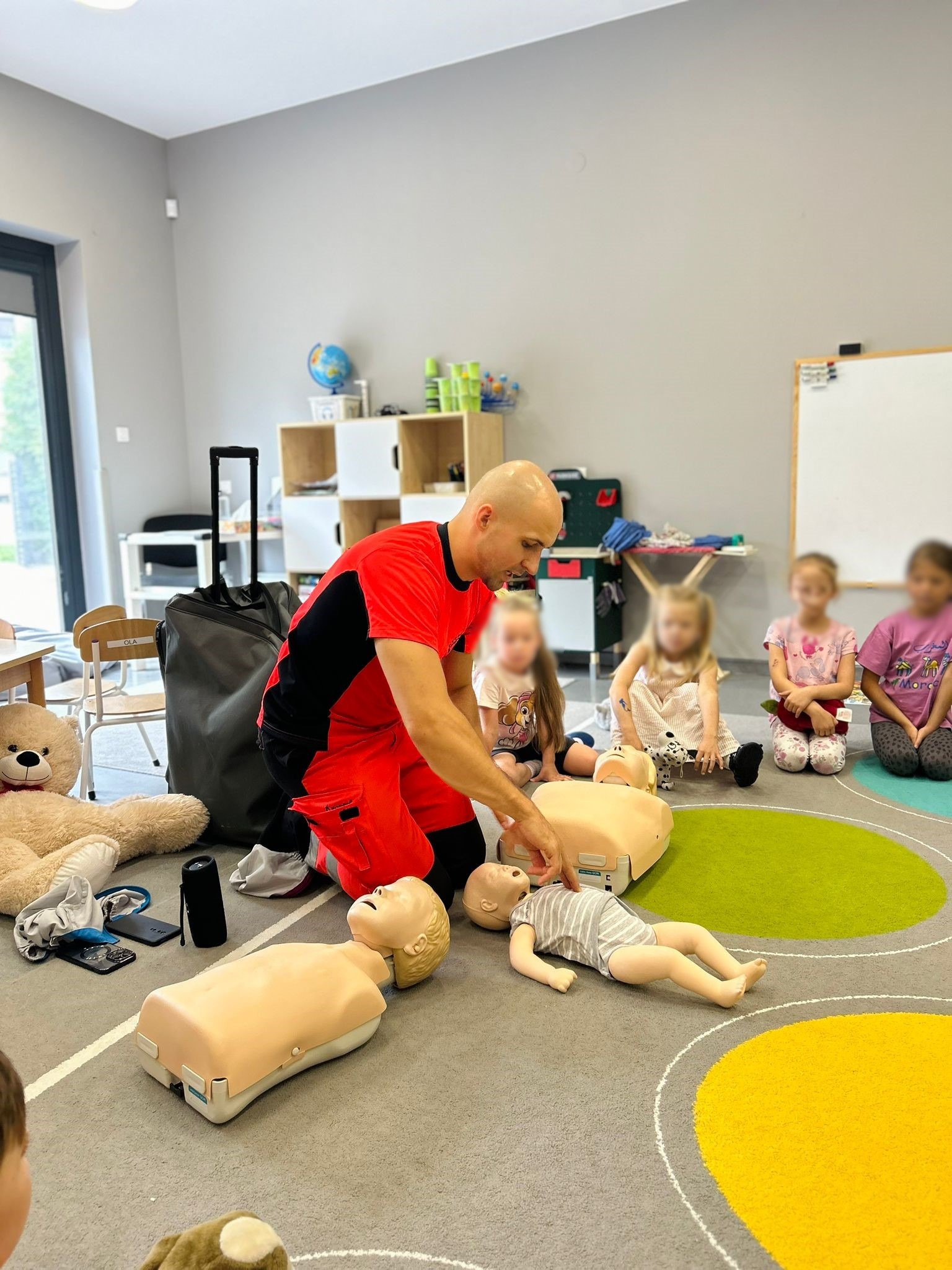 Instruktor w czerwonym uniformie demonstruje resuscytację krążeniowo-oddechową na manekinie niemowlęcia w obecności dzieci siedzących na dywanie w przedszkolu.