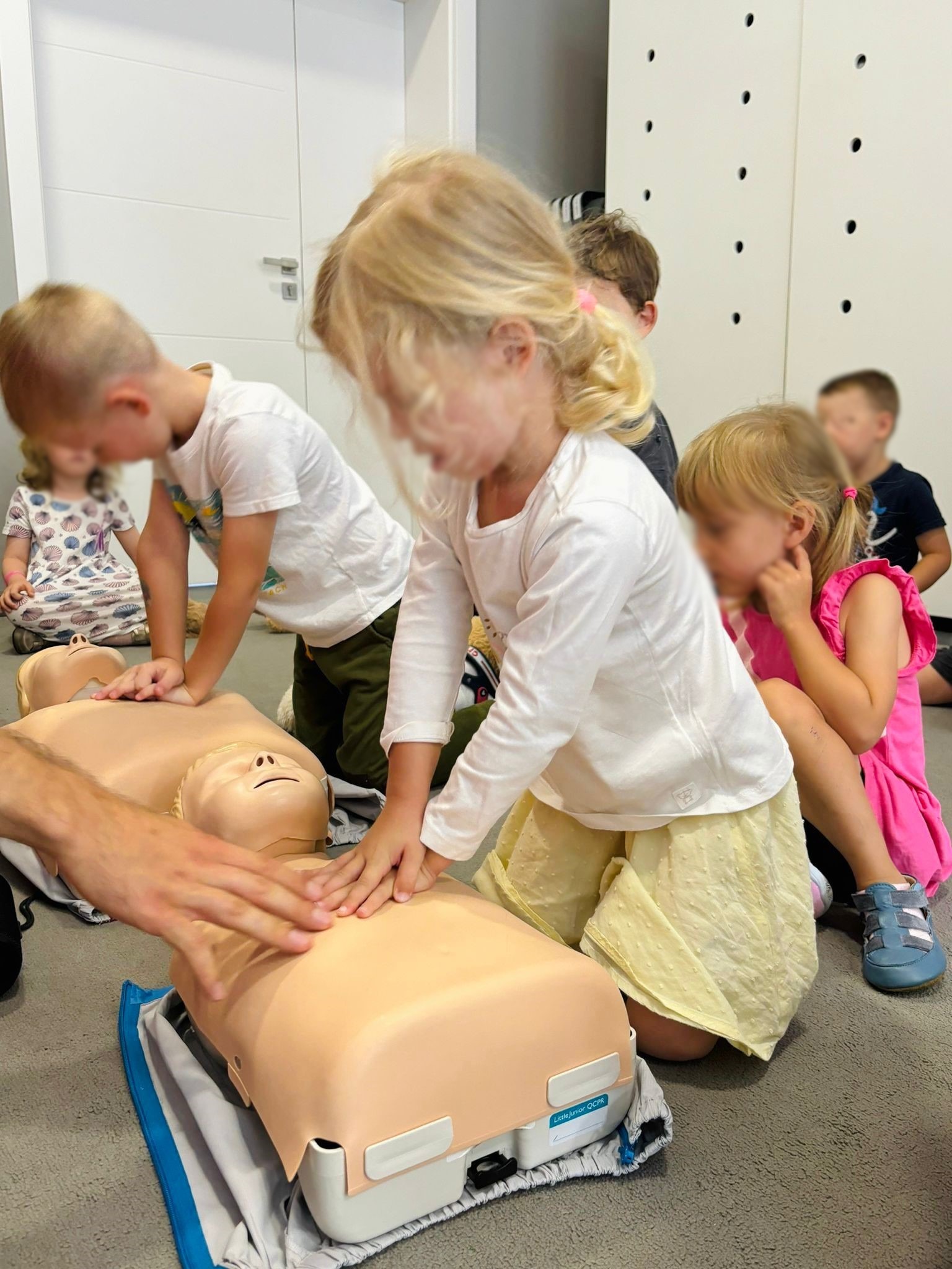 Grupa dzieci, w tym blondwłosa dziewczynka w żółtej spódnicy, uczy się resuscytacji krążeniowo-oddechowej na manekinie Little Junior QCPR pod nadzorem dorosłego, siedząc na szarym dywanie.