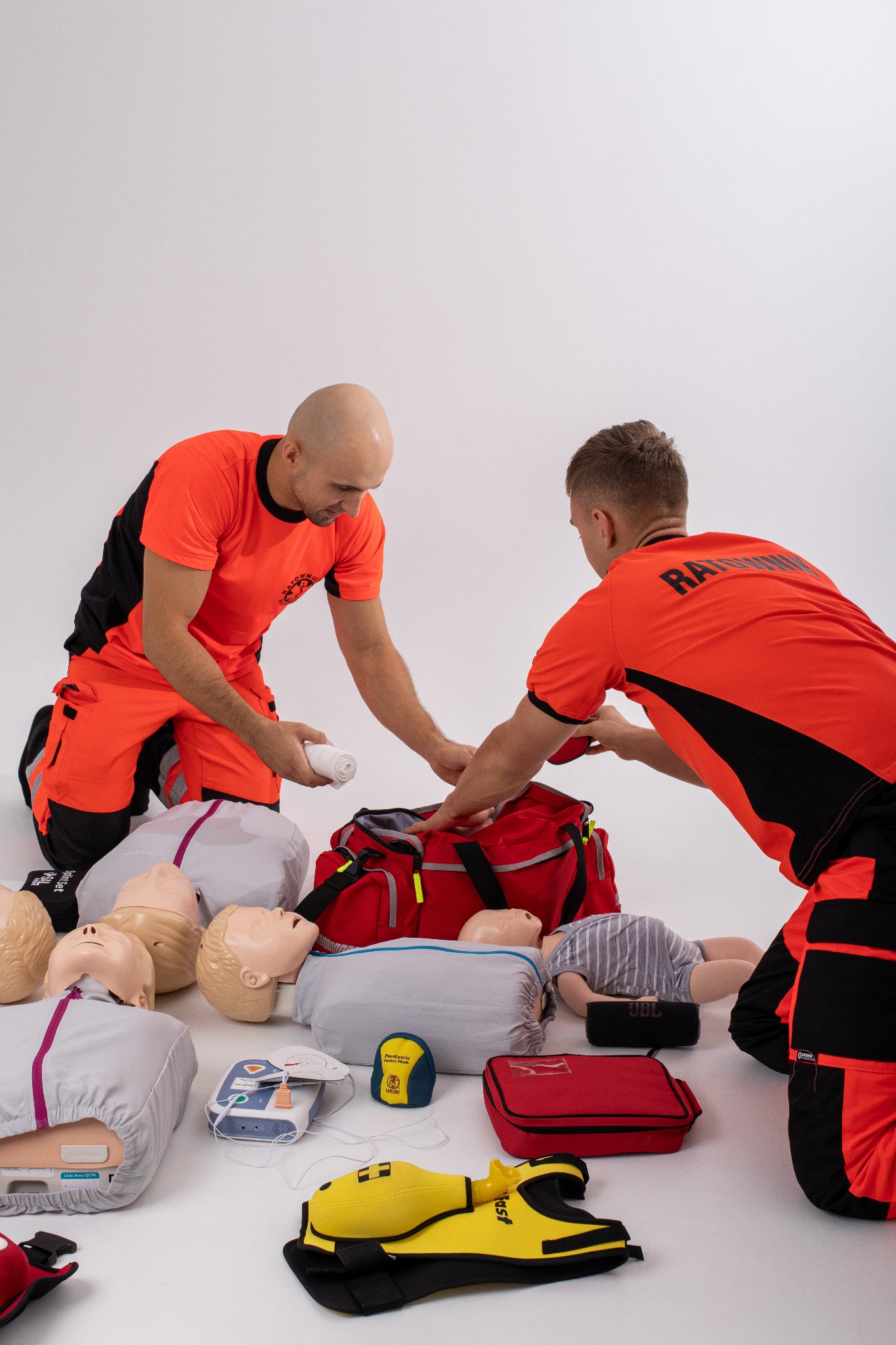 Dwóch ratowników w pomarańczowych uniformach przygotowuje torbę medyczną na białym tle, otoczoną manekinami do ćwiczeń resuscytacji, defibrylatorem i akcesoriami.