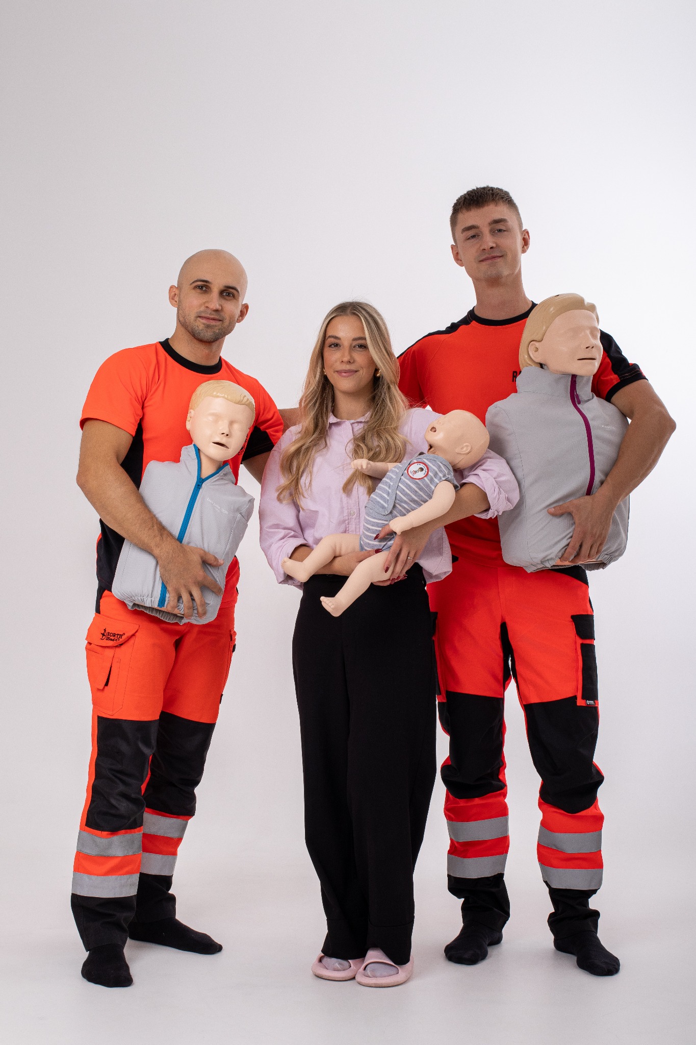 Trzy osoby pozują z manekinami do ćwiczeń resuscytacji: dwóch mężczyzn w pomarańczowo-czarnych strojach ratowniczych trzyma manekiny dorosłych, a kobieta w różowej koszuli trzyma manekin...