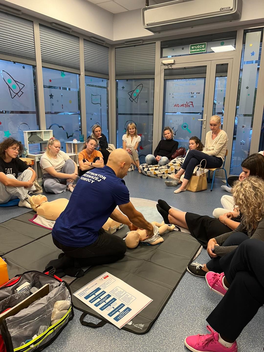 Instruktor w niebieskiej koszulce demonstruje resuscytację niemowlęcia na manekinie podczas zajęć grupowych, obok leży torba medyczna i schemat postępowania.