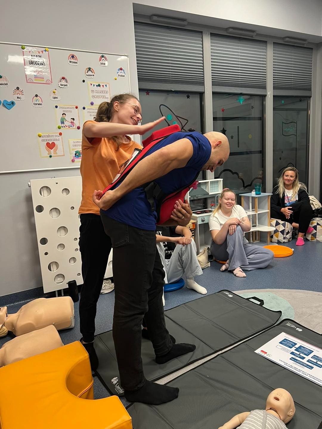 Instruktorka w pomarańczowej koszulce demonstruje na mężczyźnie w niebieskiej koszulce użycie kamizelki do ratowania osoby duszącej się, w tle manekiny i osoby obserwujące.