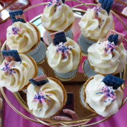 Osiem babeczek z białym kremem, posypką i czekoladowymi dekoracjami na złotym, okrągłym stojaku, na tle różowej tkaniny. W odbiciu na stojaku widać rękę trzymającą telefon.