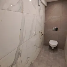 Nowoczesna łazienka z płytkami imitującymi marmur na ścianach i szarymi płytkami podłogowymi. Widoczna toaleta i chromowane uchwyty dla niepełnosprawnych.