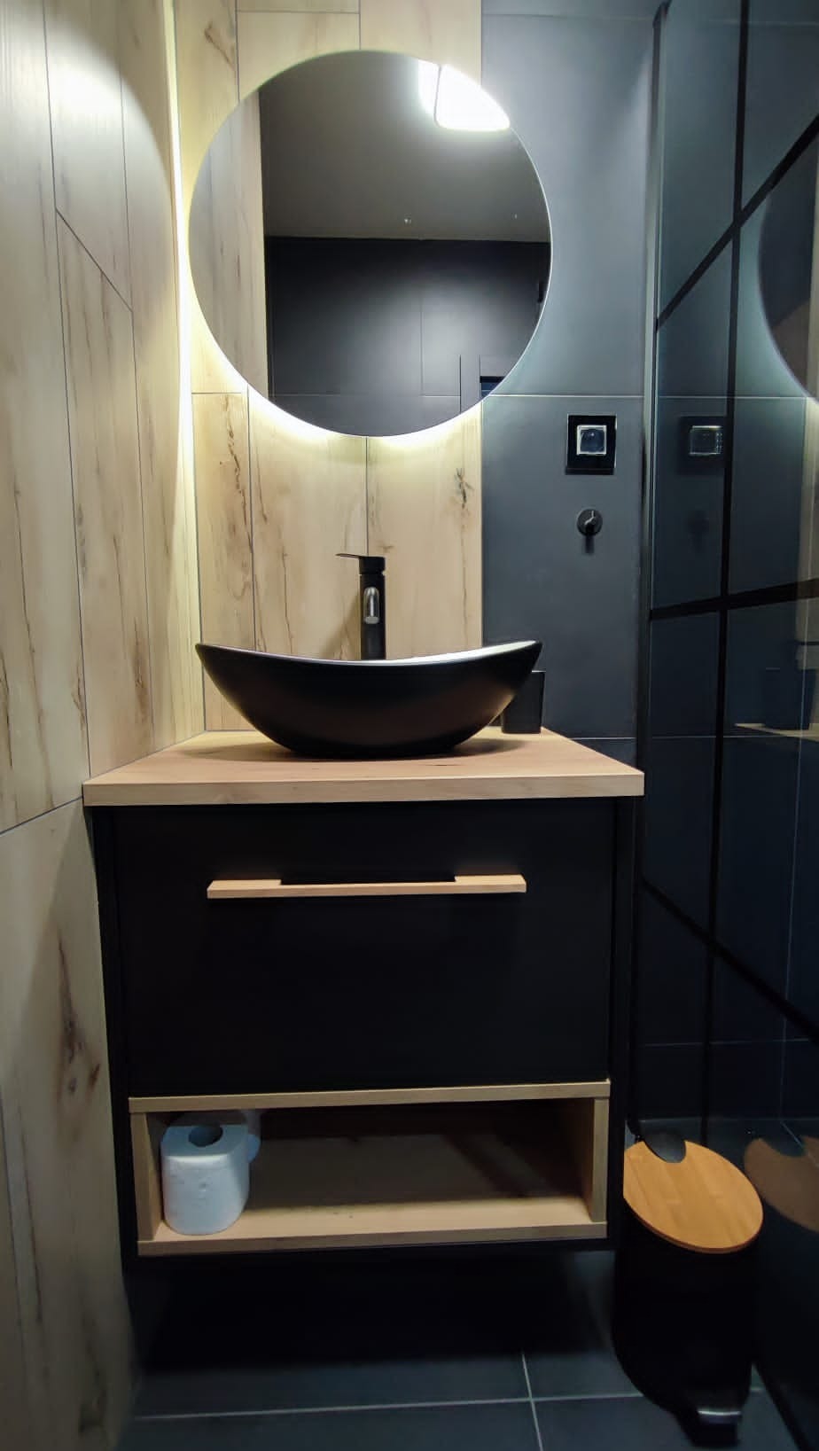 Nowoczesna łazienka z czarną umywalką na drewnianej szafce, okrągłe lustro z podświetleniem LED i grafitowe płytki. Minimalistyczny design z akcentami drewna.