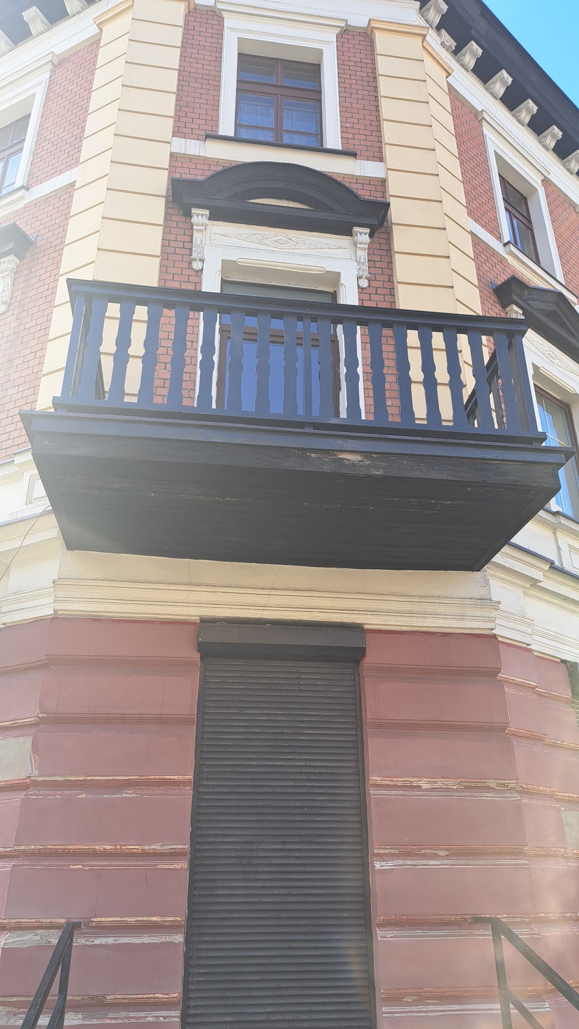 Elewacja kamienicy z czarnym, odnowionym balkonem z ozdobną balustradą, widok z dołu. Zamknięta roleta na parterze. Czerwona cegła i kremowe zdobienia.